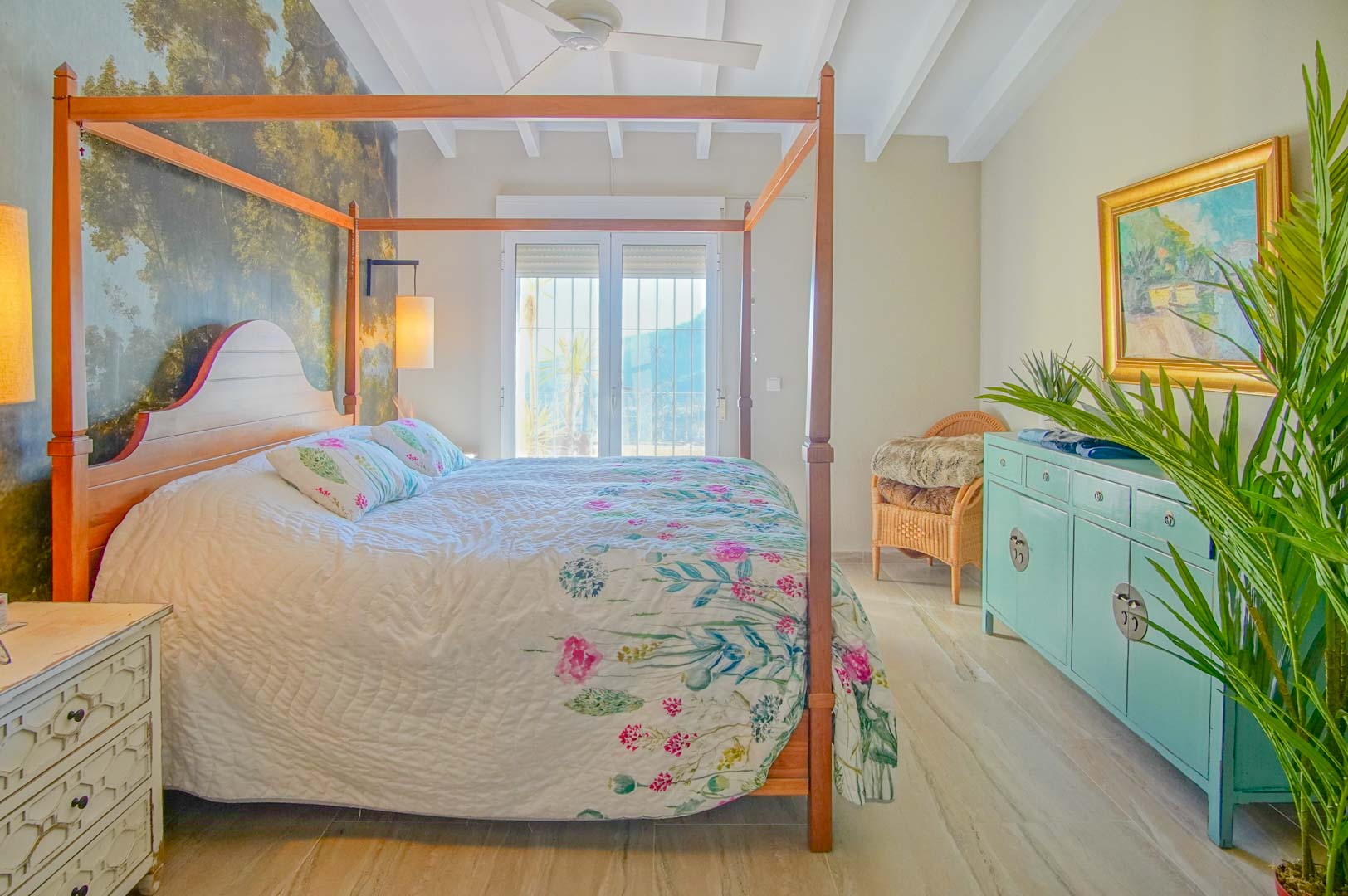 Bedroom featuring a wooden four-poster bed, forest wall mural, white beamed ceiling with fan, and balcony access providing abundant natural light.