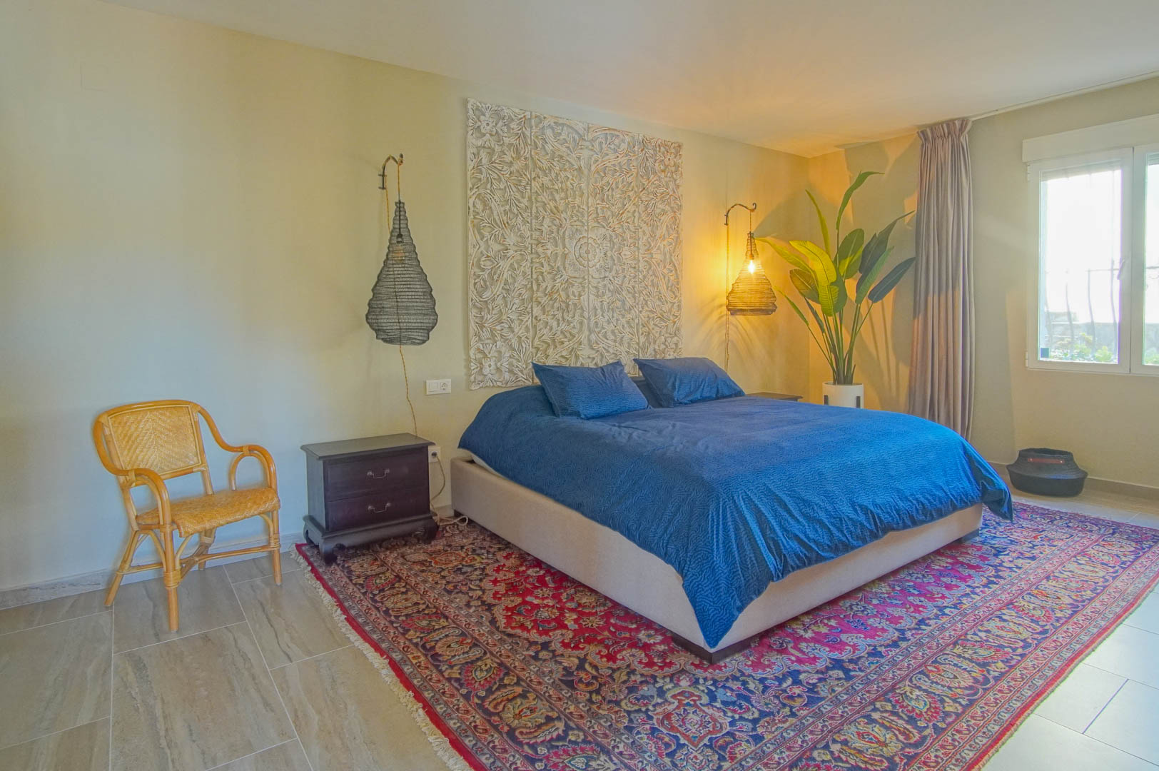 Bedroom featuring an ornate carved wall panel, woven pendant lights, and a large oriental rug over tiled flooring. Natural light enters through a side window.