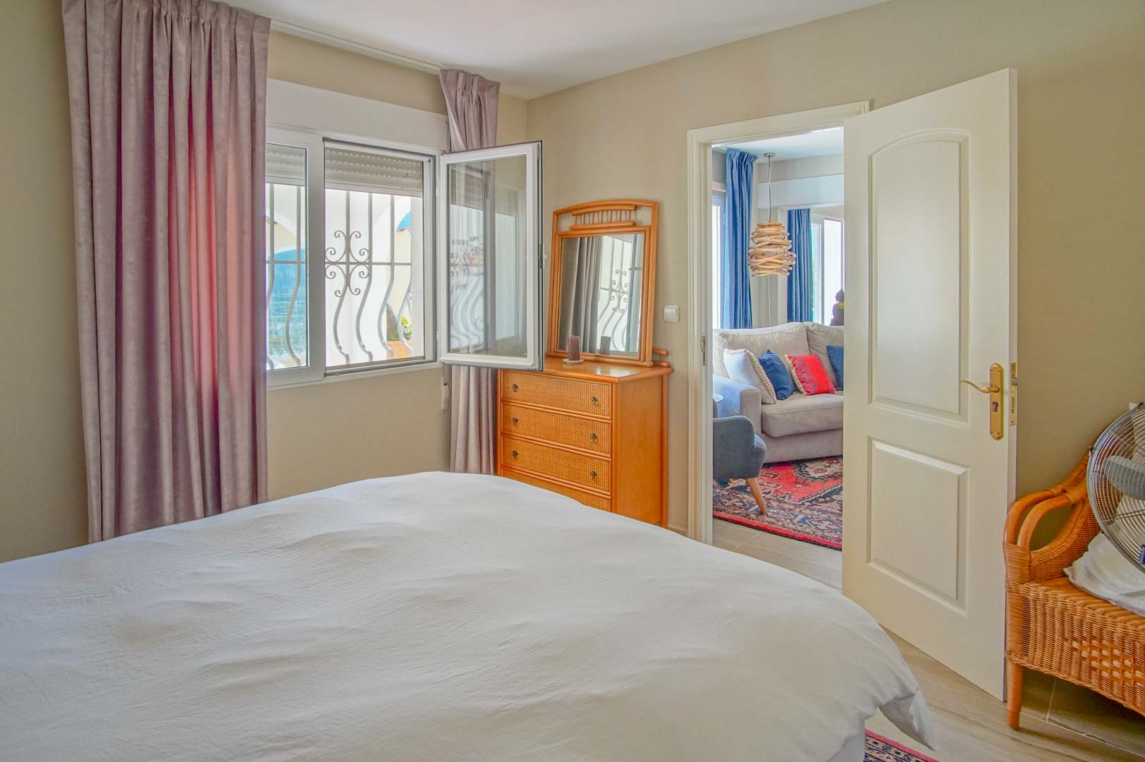 Bright bedroom featuring a wooden dresser and decorative window grilles. An open doorway connects to an adjacent living area with a sofa and patterned rug.
