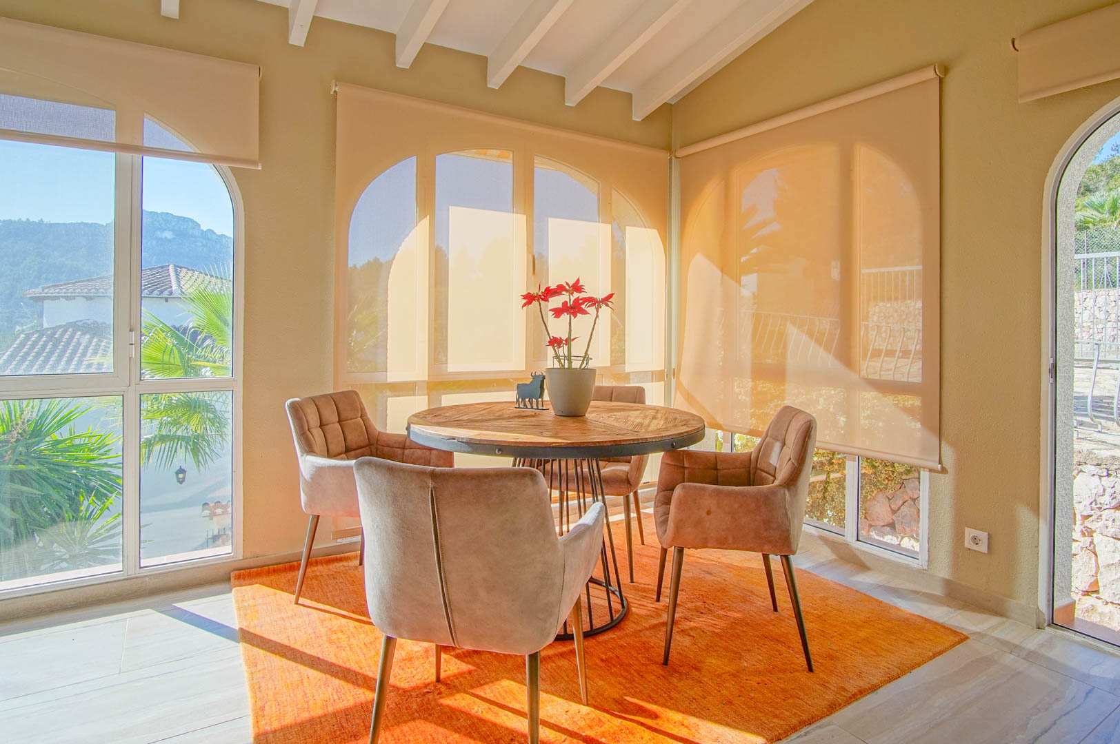 Bright dining space featuring a round wooden table, exposed white ceiling beams, and large windows with mountain views and roller blinds.