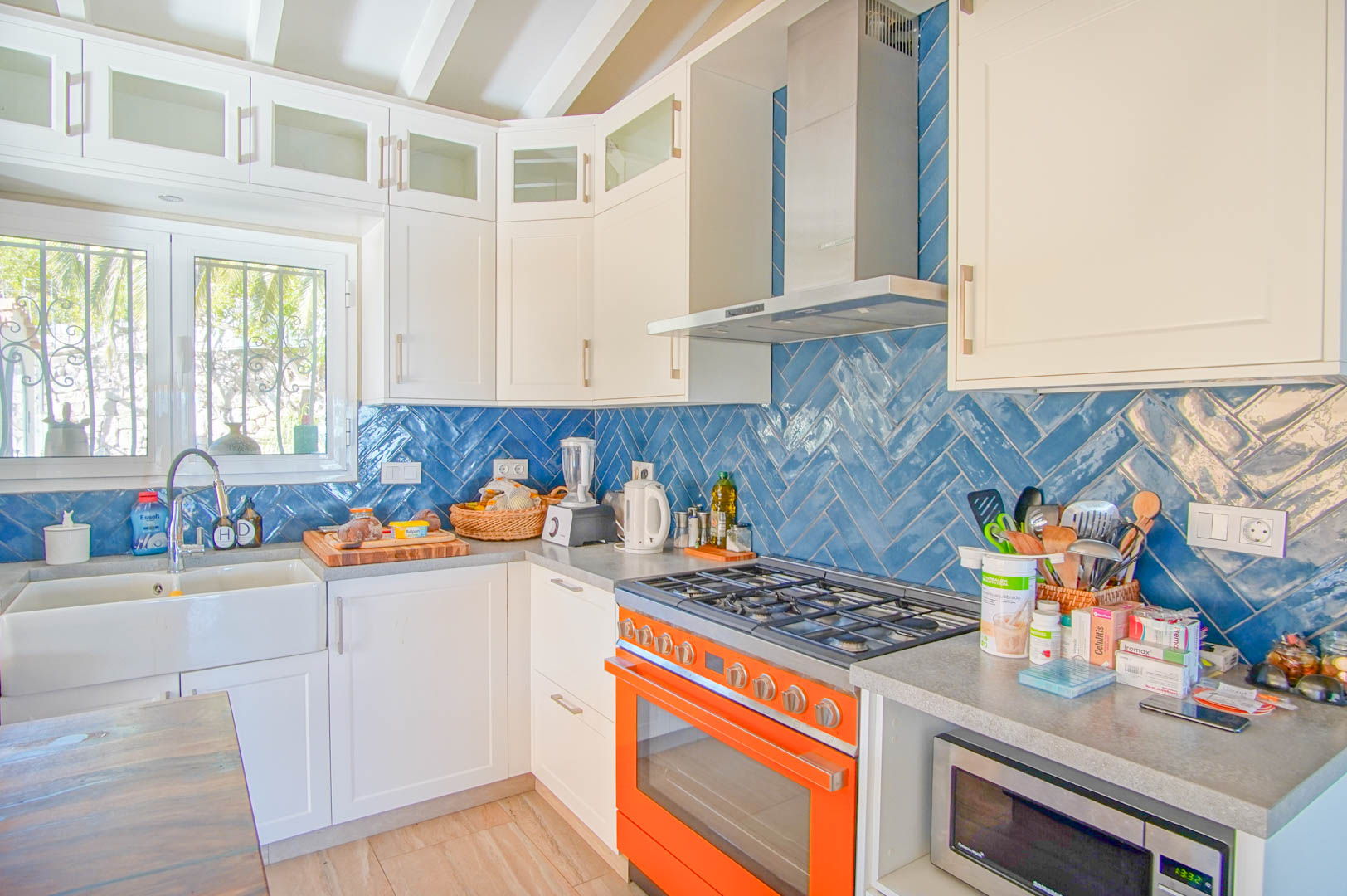 Bright kitchen featuring a blue herringbone tile backsplash, grey countertops, and a bold orange range. It includes a farmhouse sink and white exposed ceiling beams.