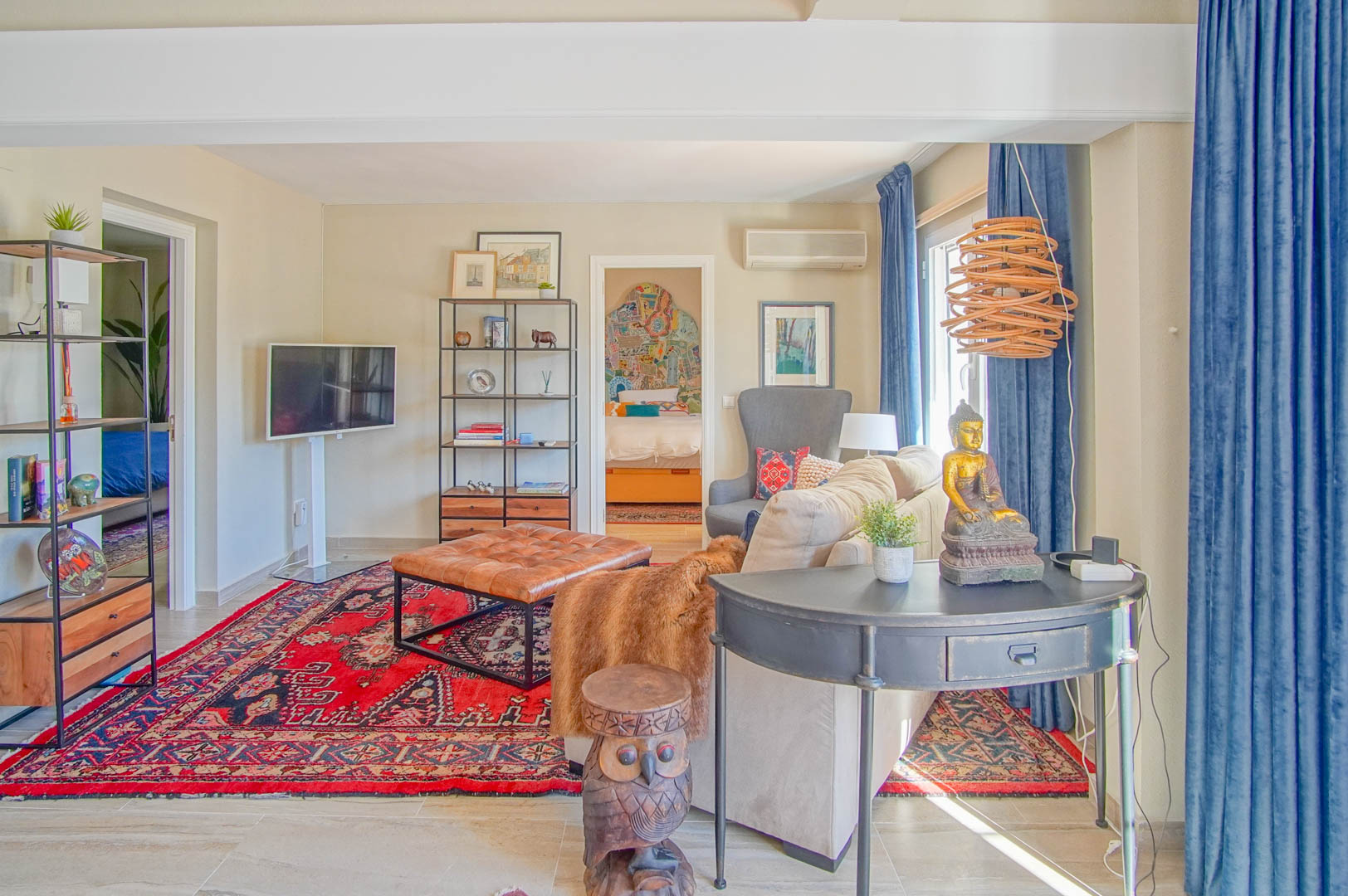 Bright living area featuring a red oriental rug, tufted leather ottoman, and eclectic decor, with a view into an adjacent bedroom through an open doorway.