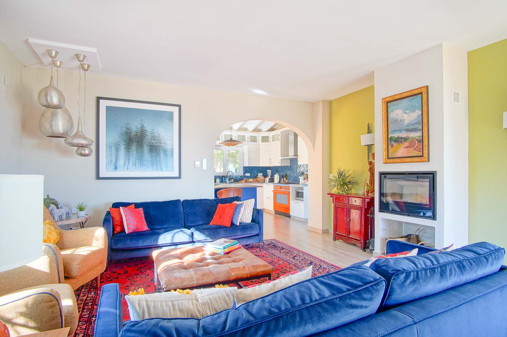 Bright living area featuring blue velvet sofas, a built-in fireplace, and an archway to the kitchen. It includes silver pendant lights and light wood-effect flooring.