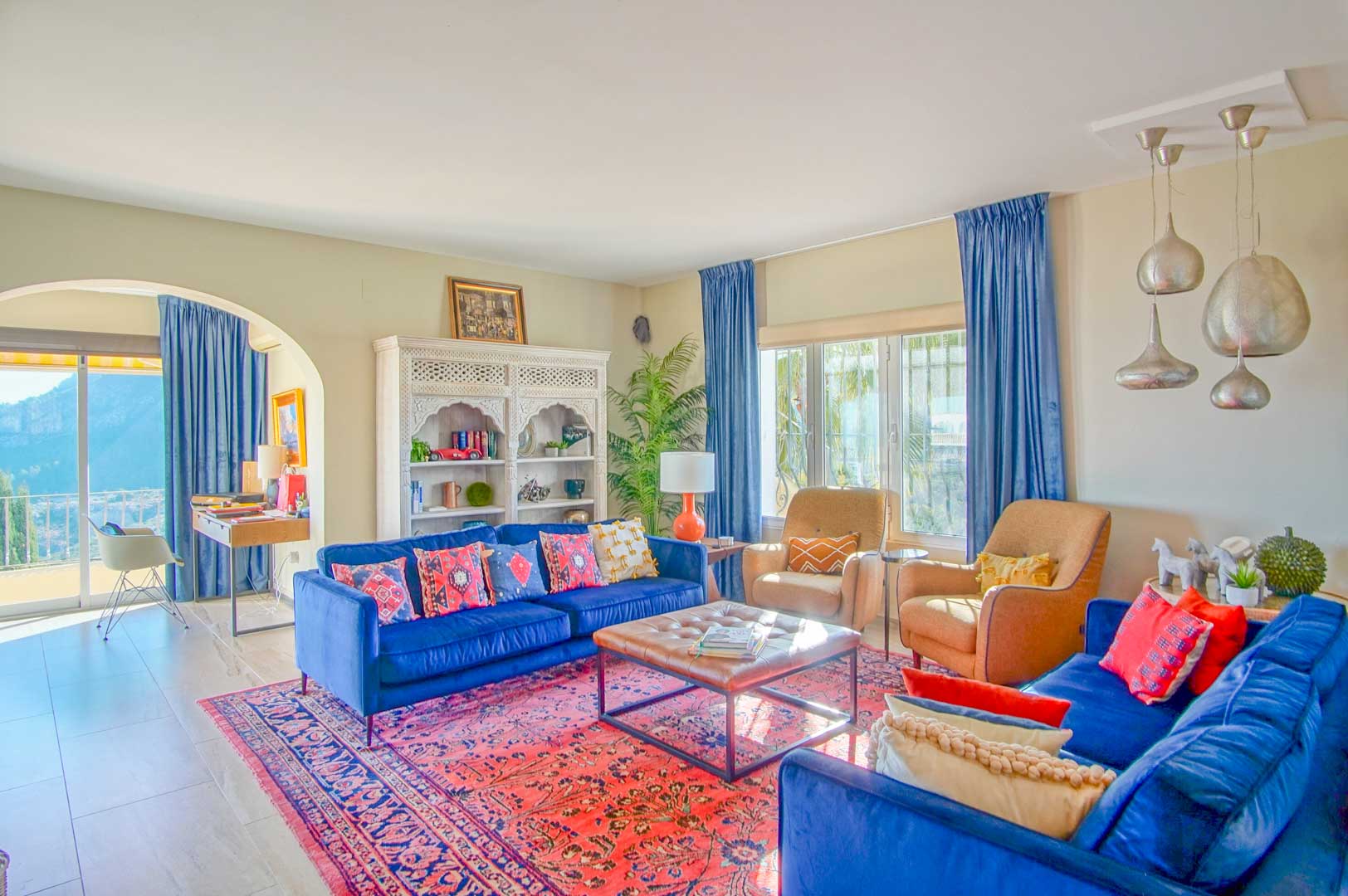 Bright living area featuring blue velvet sofas, an oriental rug, and a carved white cabinet. An archway leads to a workspace with mountain views.