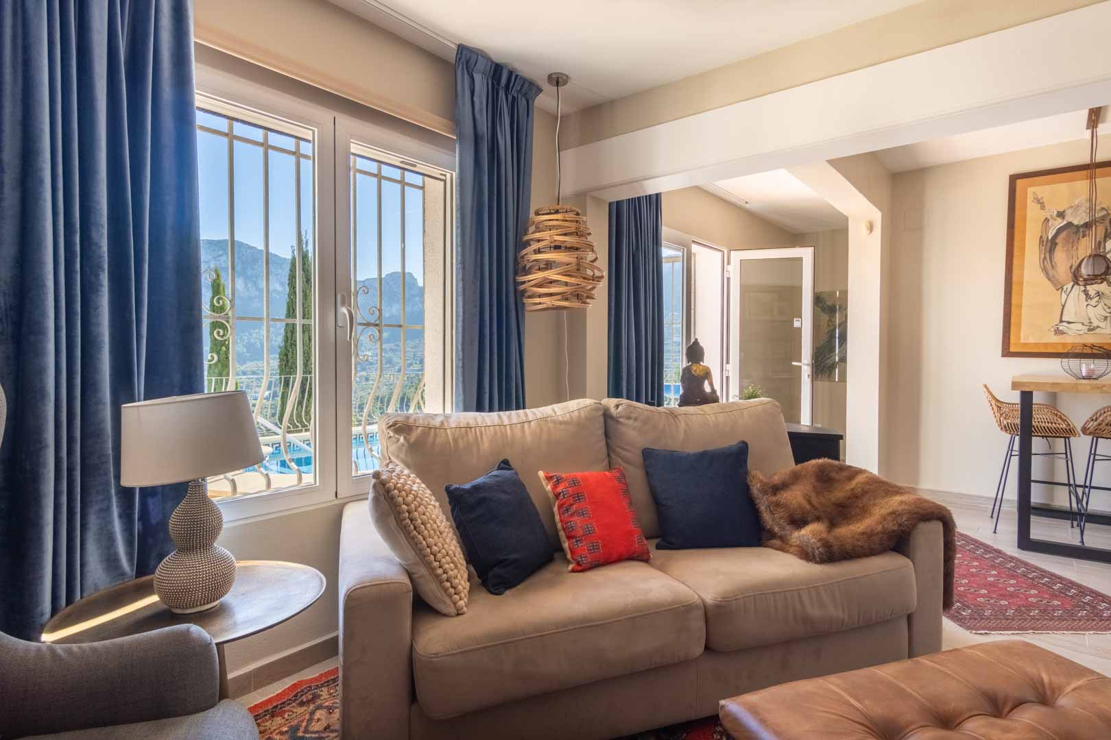 Bright living area featuring earth-toned seating, blue velvet curtains, and large windows overlooking the mountains. An adjacent breakfast bar and ethnic decor complete the space.