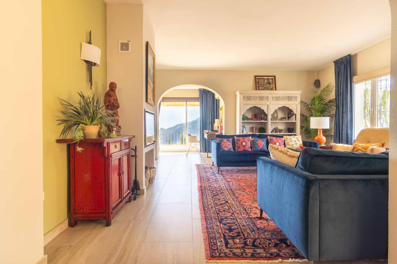 Bright living room featuring blue velvet sofas, an oriental rug, and an arched doorway framing mountain views. Includes a carved white cabinet and tiled flooring.