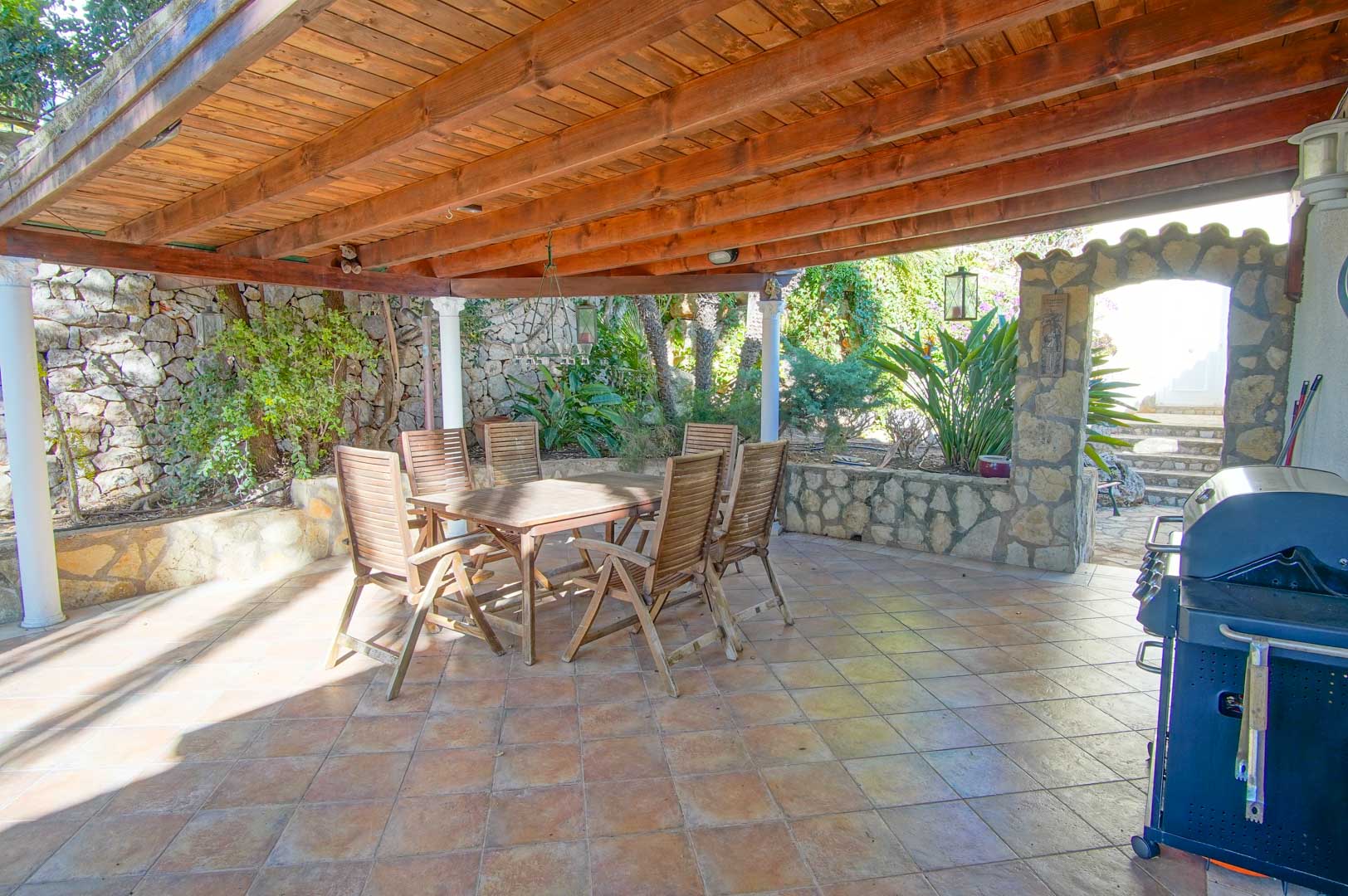 Covered outdoor dining area featuring a robust wooden pergola, stone masonry walls, a barbecue grill, and a rustic wooden table set.