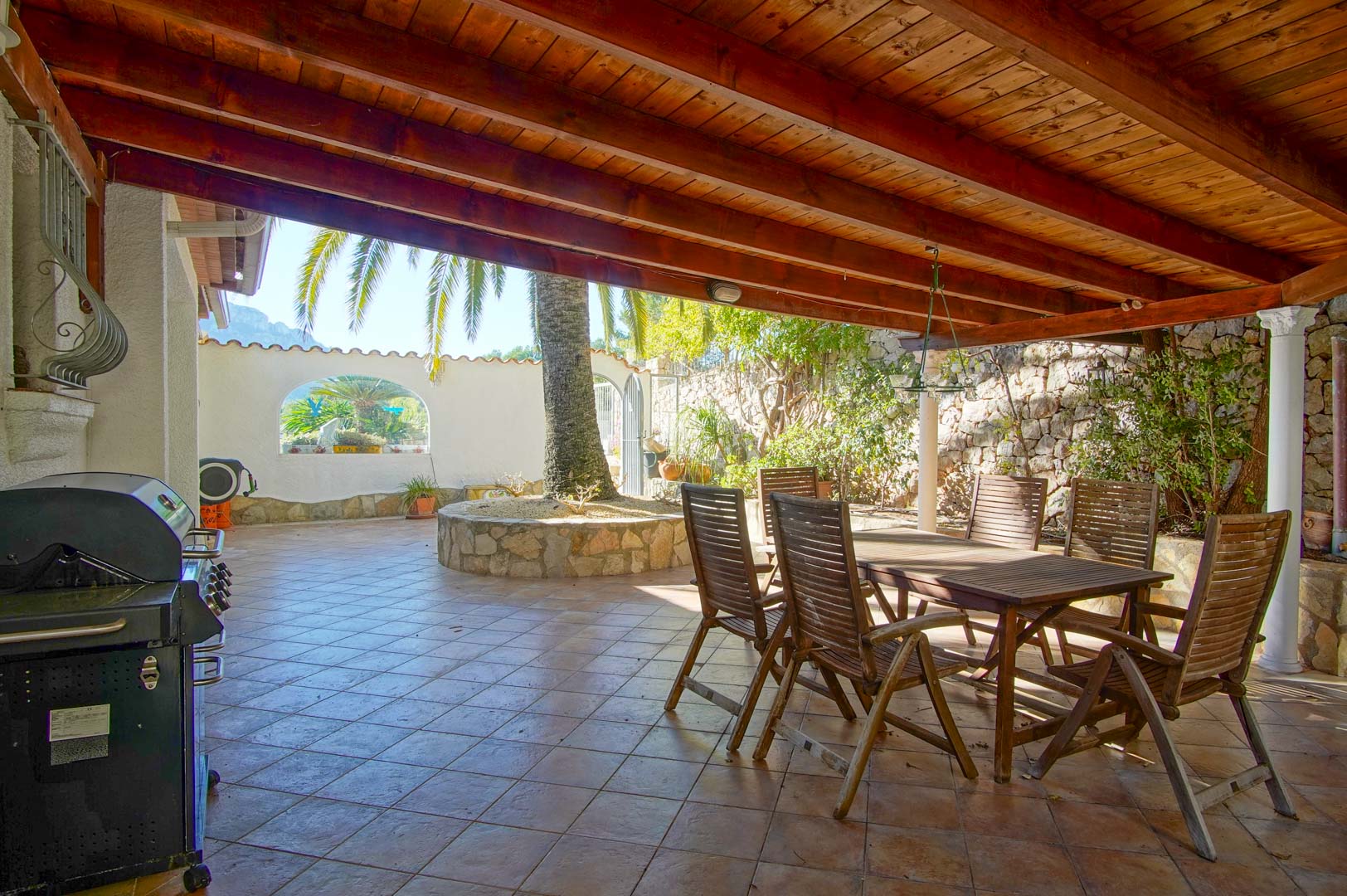 Covered terrace featuring a wooden beam ceiling, outdoor dining set, gas barbecue, and a circular stone planter with a palm tree against natural stone walls.