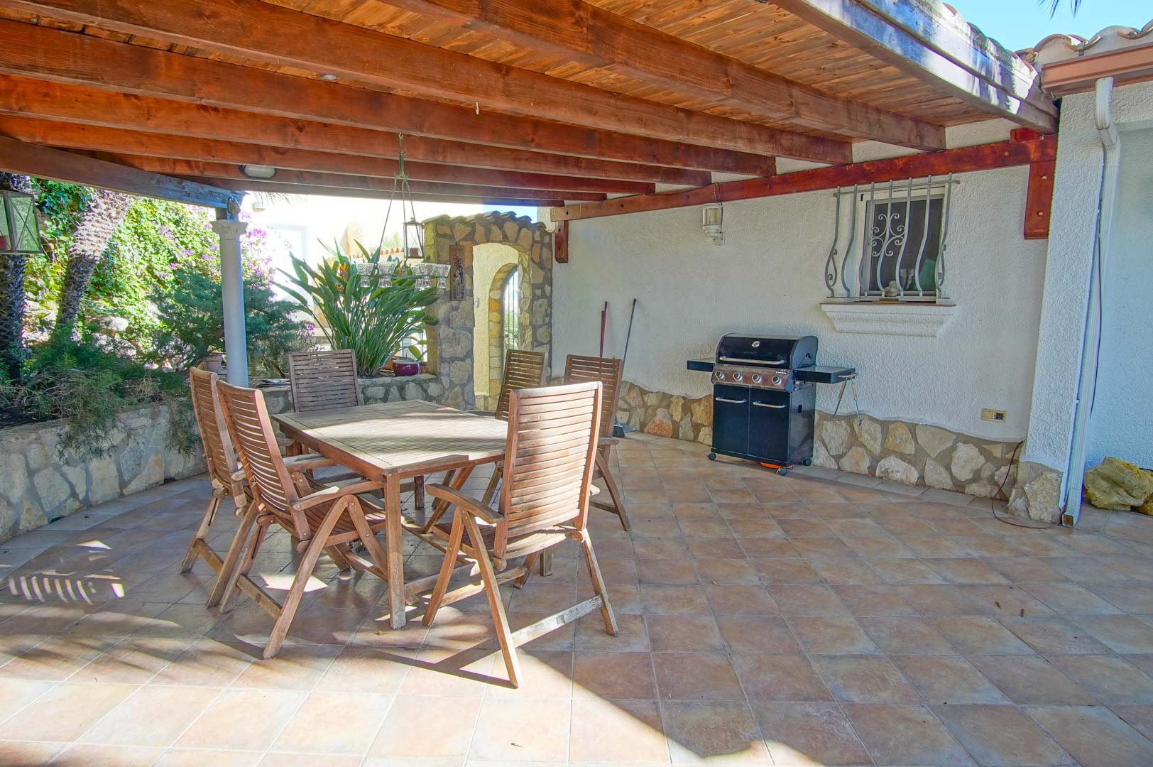 Covered terrace featuring a wooden pergola, outdoor dining set, and gas barbecue grill set against stone-clad walls and tiled flooring.