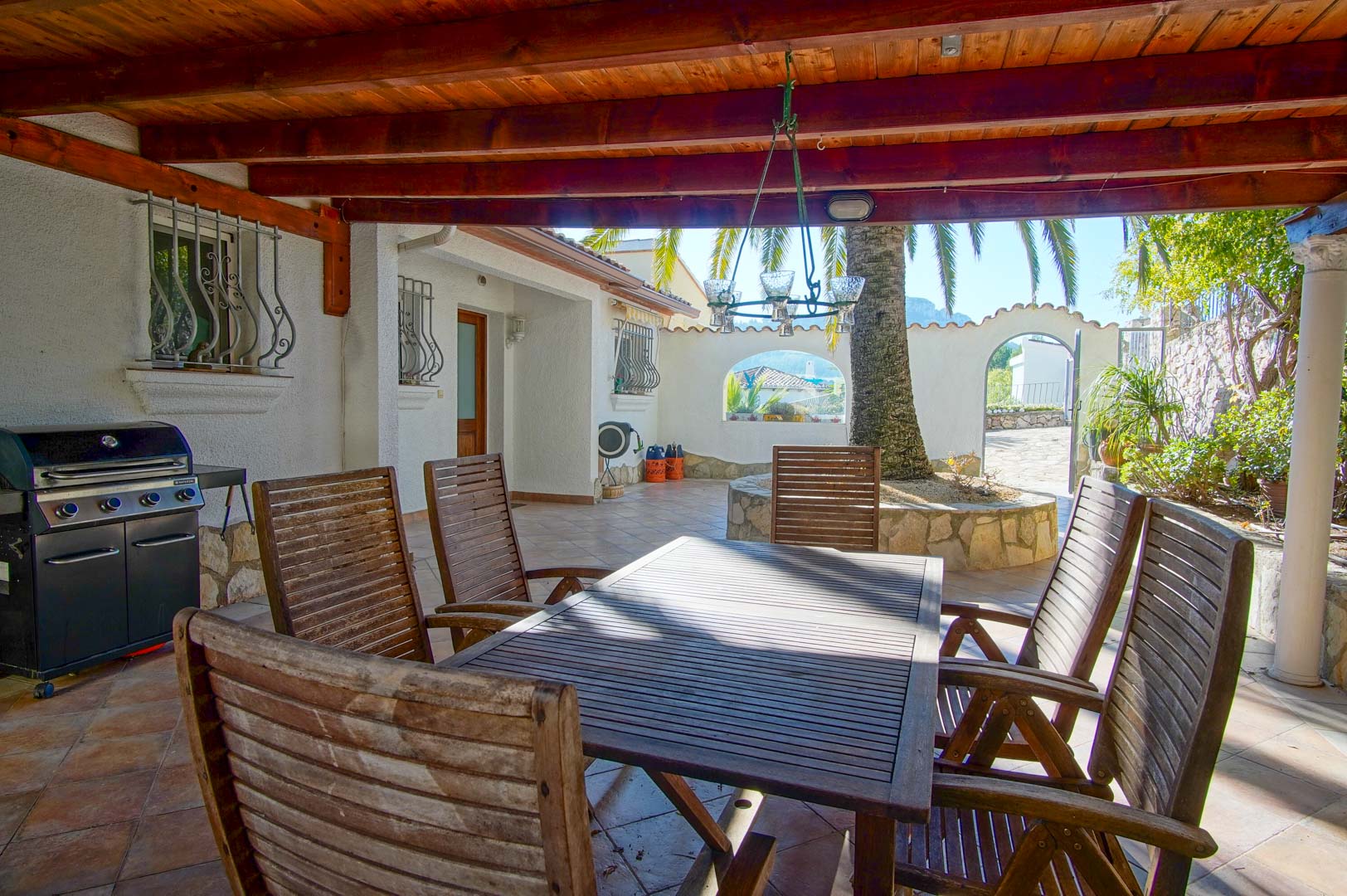 Covered terrace with a wooden beam ceiling, outdoor dining area, gas barbecue, and a courtyard featuring a palm tree and stone masonry.