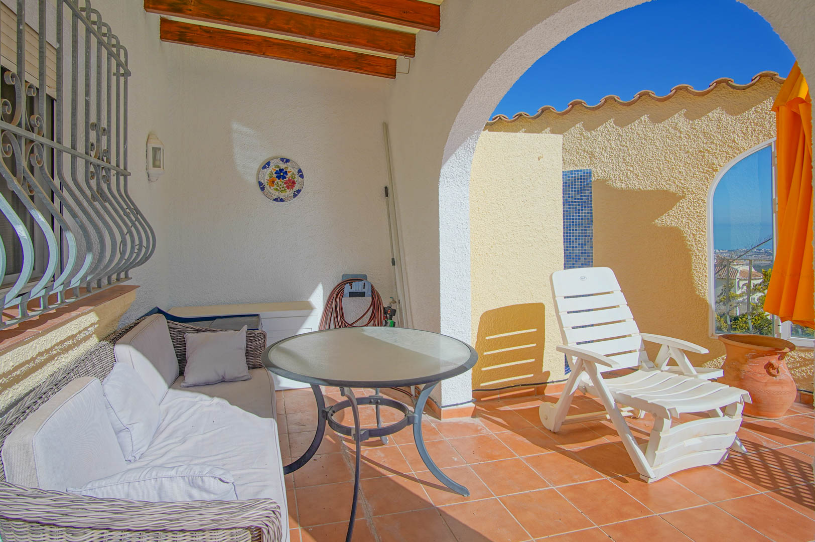 Covered terrace with exposed wooden beams, terracotta tiles, and lounge furniture leading to a sun deck with sea views and a mosaic shower.