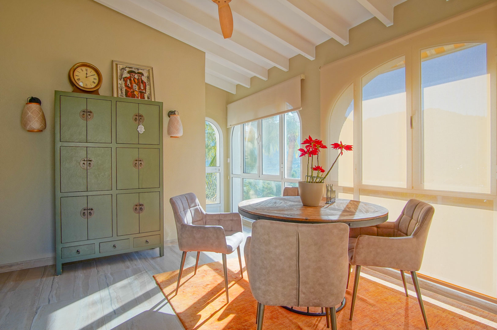 Dining area featuring a round wooden table, upholstered chairs, and white beamed ceilings. Large arched windows fill the space with natural light and offer garden views.