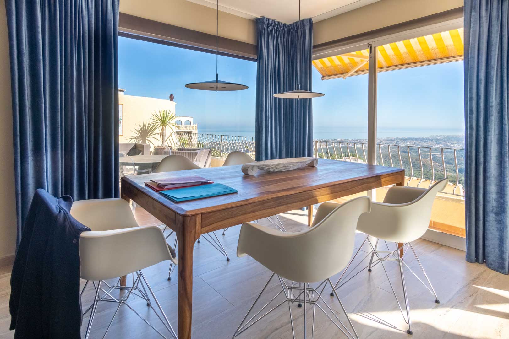Dining area featuring a solid wood table and designer chairs, framed by floor-to-ceiling windows with blue curtains and expansive Mediterranean views.