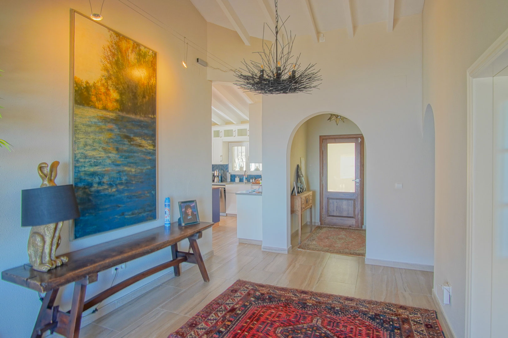 Entrance hall featuring high vaulted ceilings with white beams and an arched transition to the kitchen. Includes a rustic wooden console and a unique branch-style chandelier.