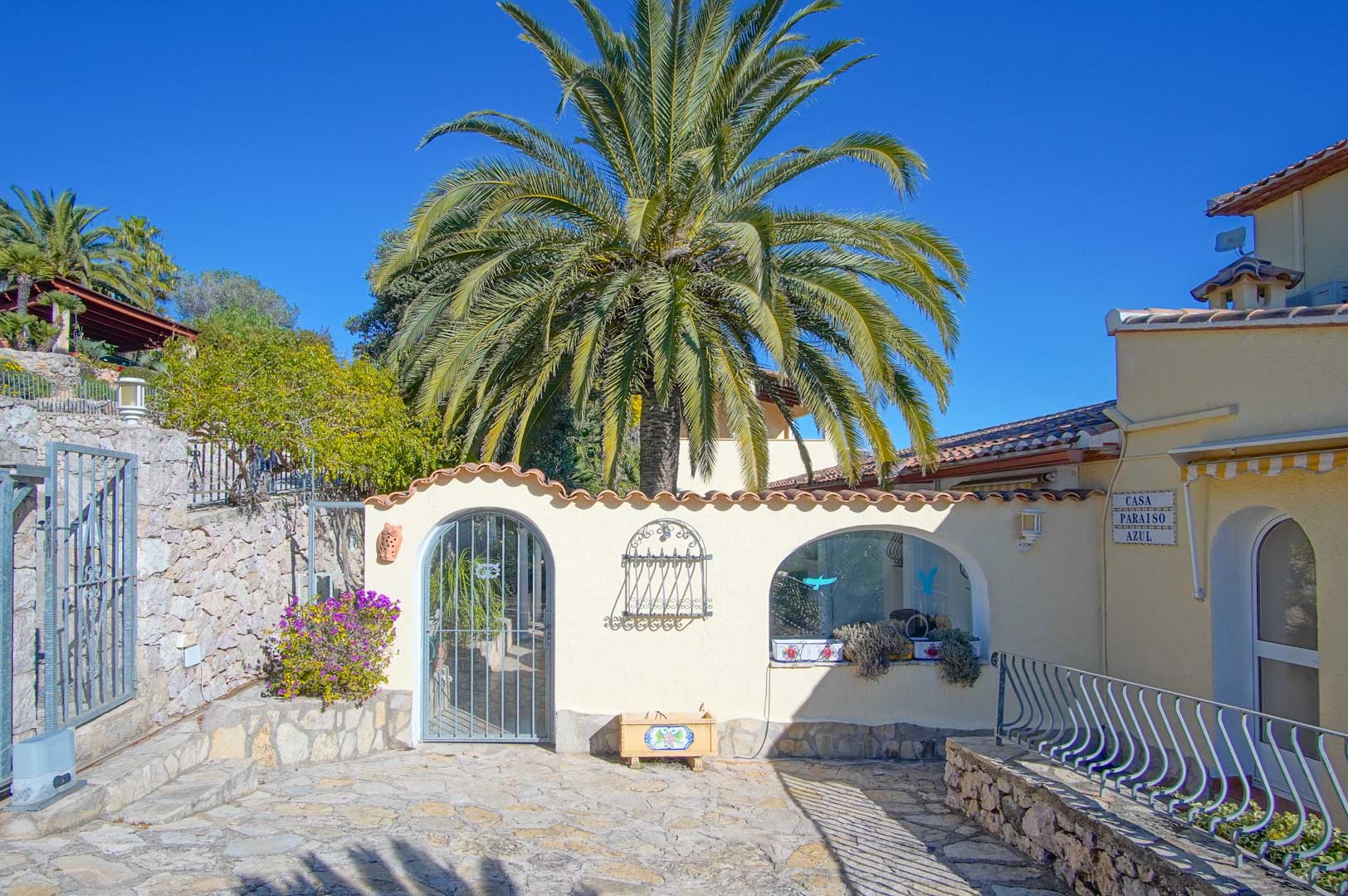 Exterior entrance featuring natural stone paving, white walls with terracotta tile trim, and a large central palm tree under a clear sky.