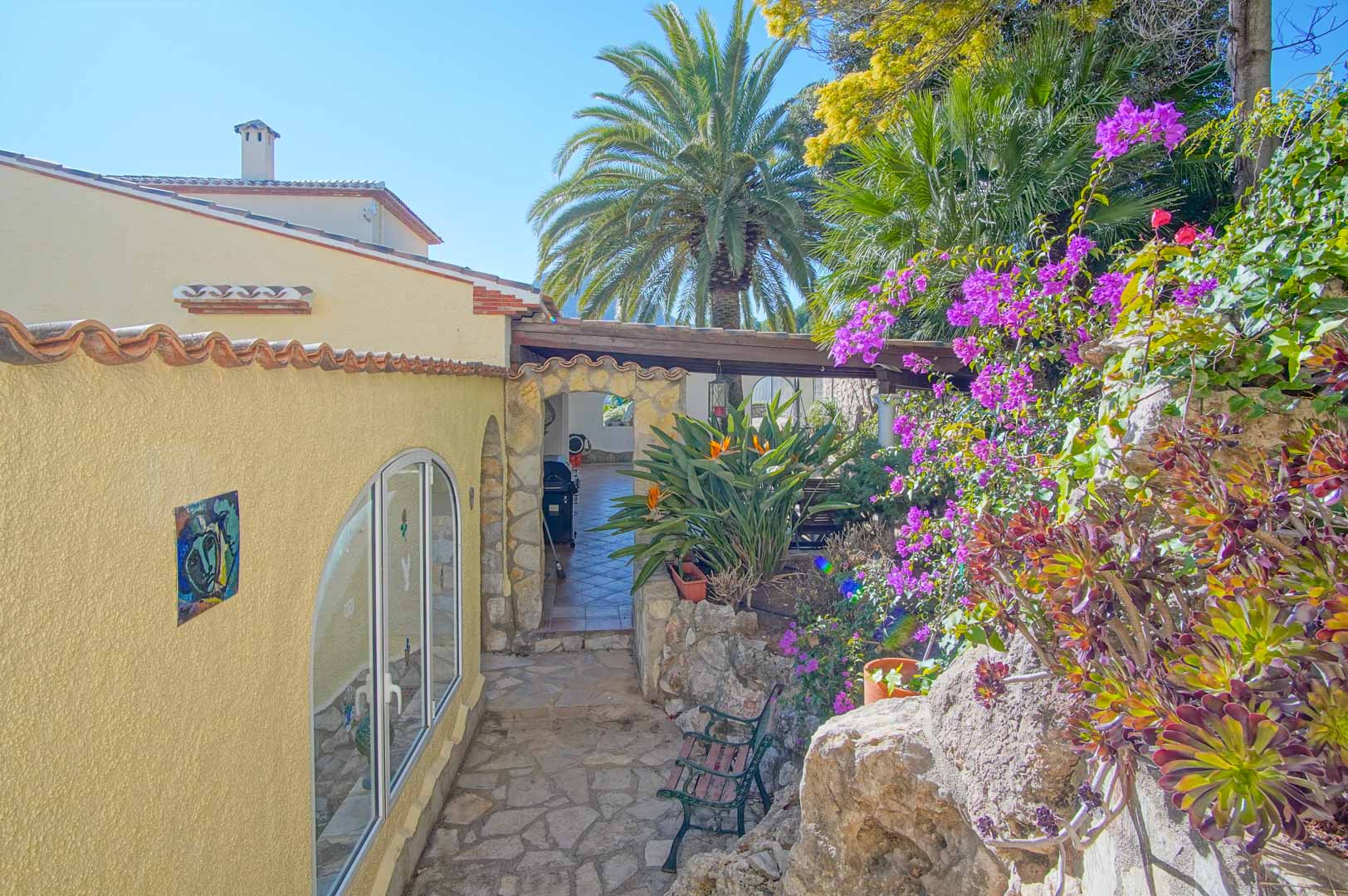 Exterior stone pathway lined with vibrant bougainvillea and Mediterranean plants, leading through a rustic stone archway to a shaded terrace area.