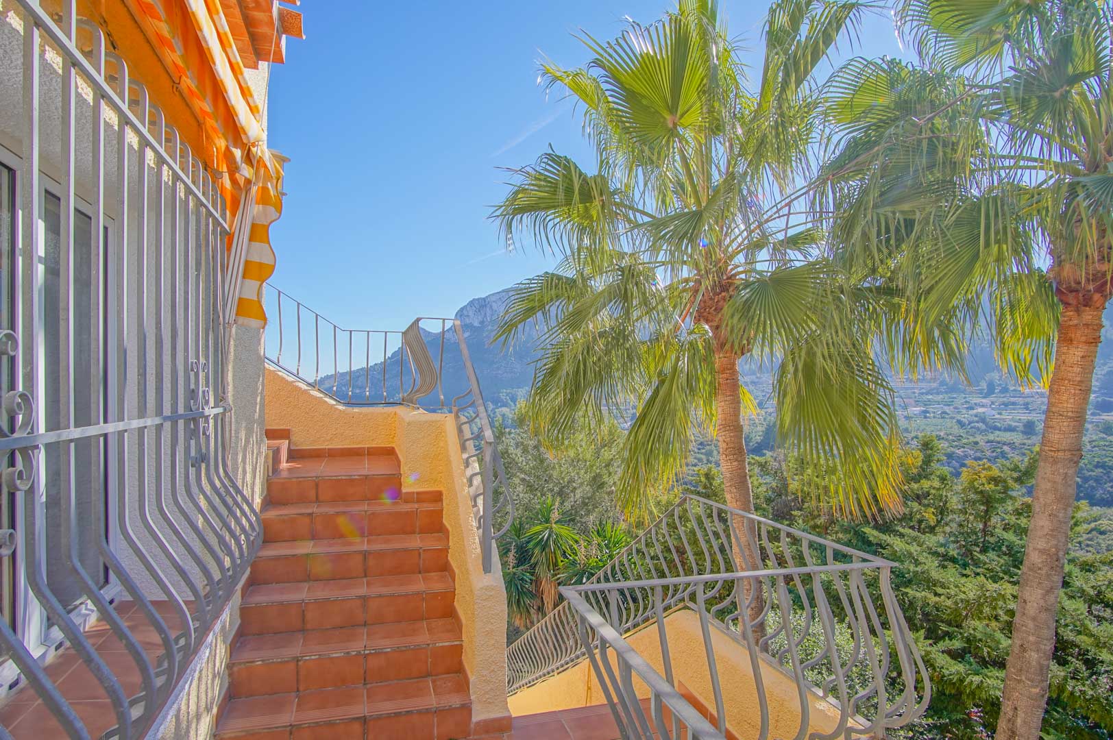Exterior terracotta staircase with wrought iron railings, featuring mountain views and mature palm trees within the Mediterranean landscape.