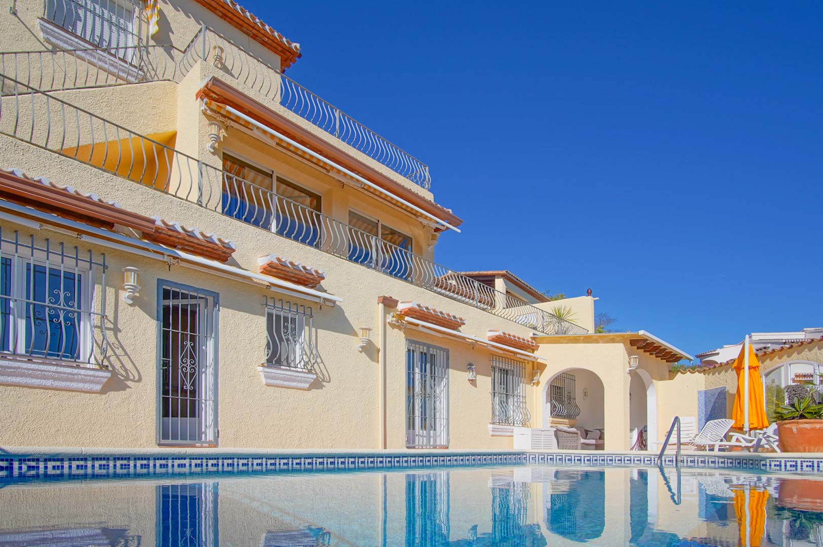 Exterior view from the pool level highlighting the three-story facade with wrought-iron terraces, sun deck with loungers, and shaded chill-out area.