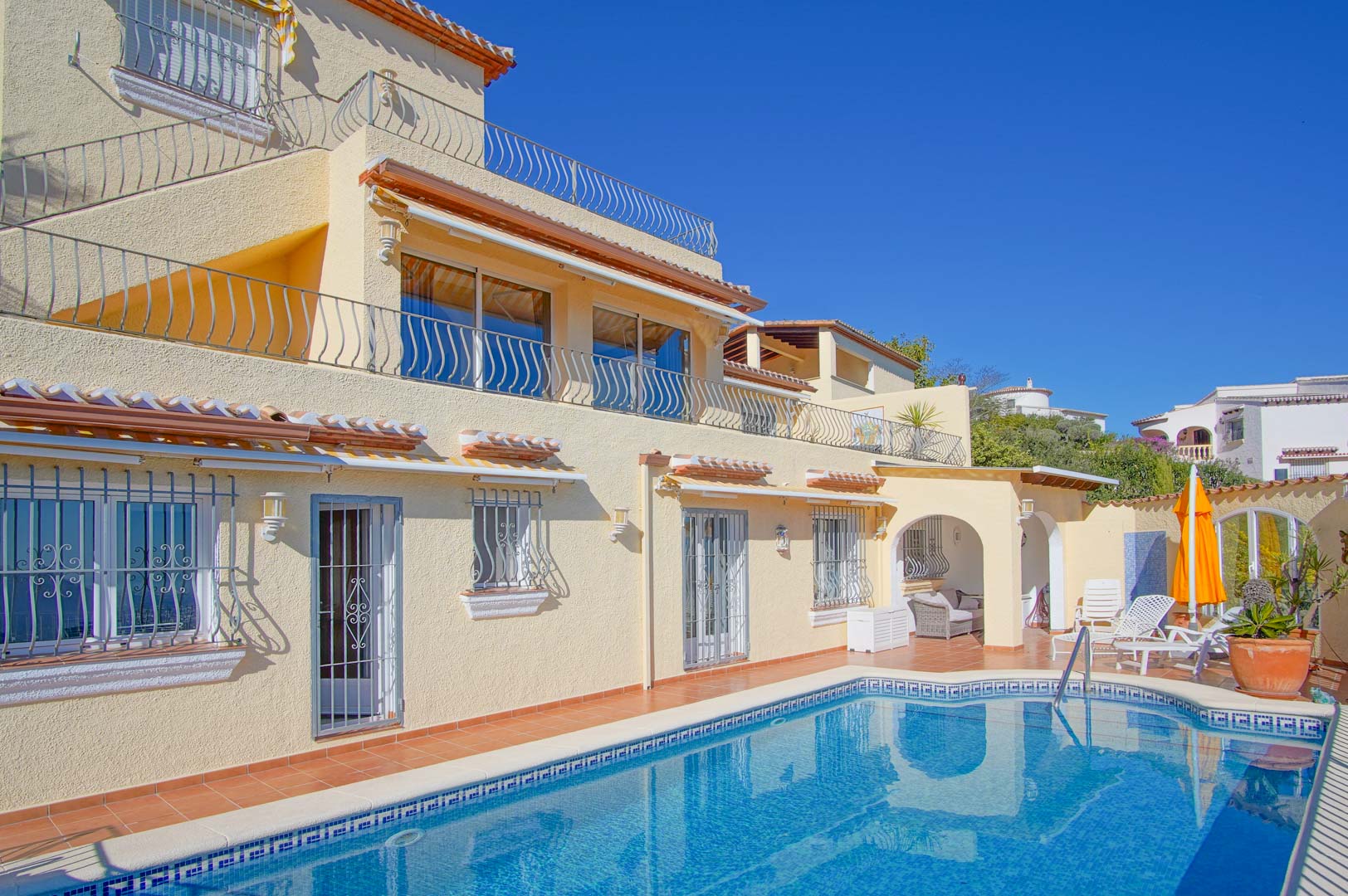 Exterior view of the villa featuring a private swimming pool, terracotta terraces, and a three-level facade. It includes an arched chill-out area and wrought iron railings.
