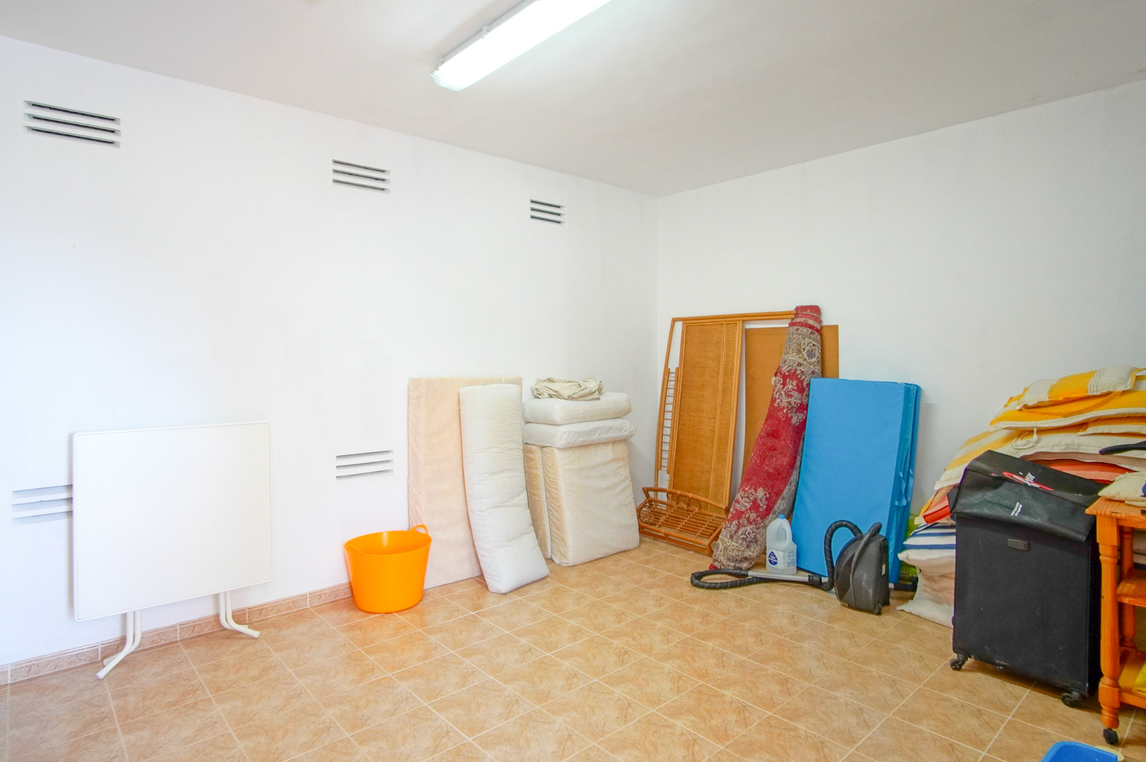 Functional storage room featuring white walls with ventilation slats and ceramic tile flooring. A versatile space with fluorescent lighting, suitable for utility needs.