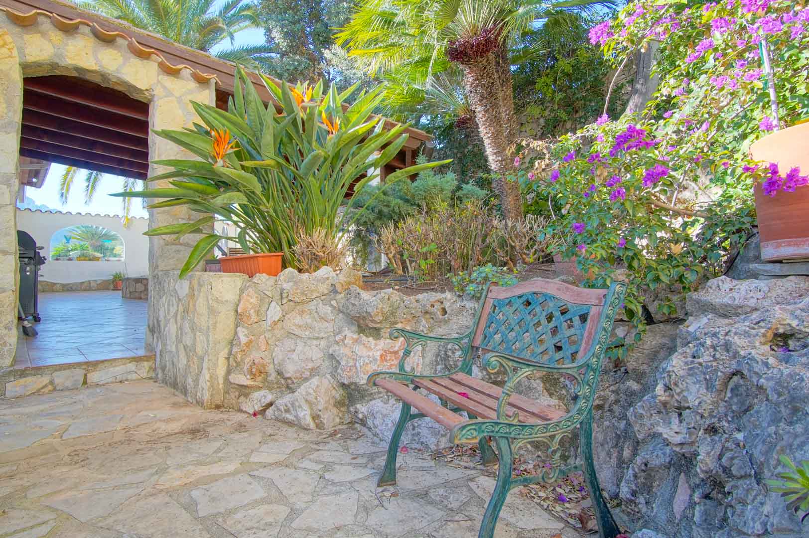 Garden seating area with a classic bench and natural stone walls, surrounded by Mediterranean flora and leading to a covered terrace with wooden beams.