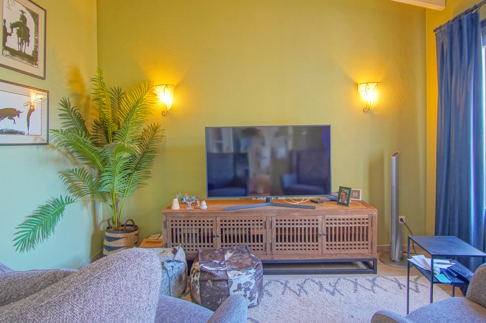 Living area featuring a wooden media console with lattice doors, warm wall sconce lighting, and a cozy seating arrangement with patterned poufs.