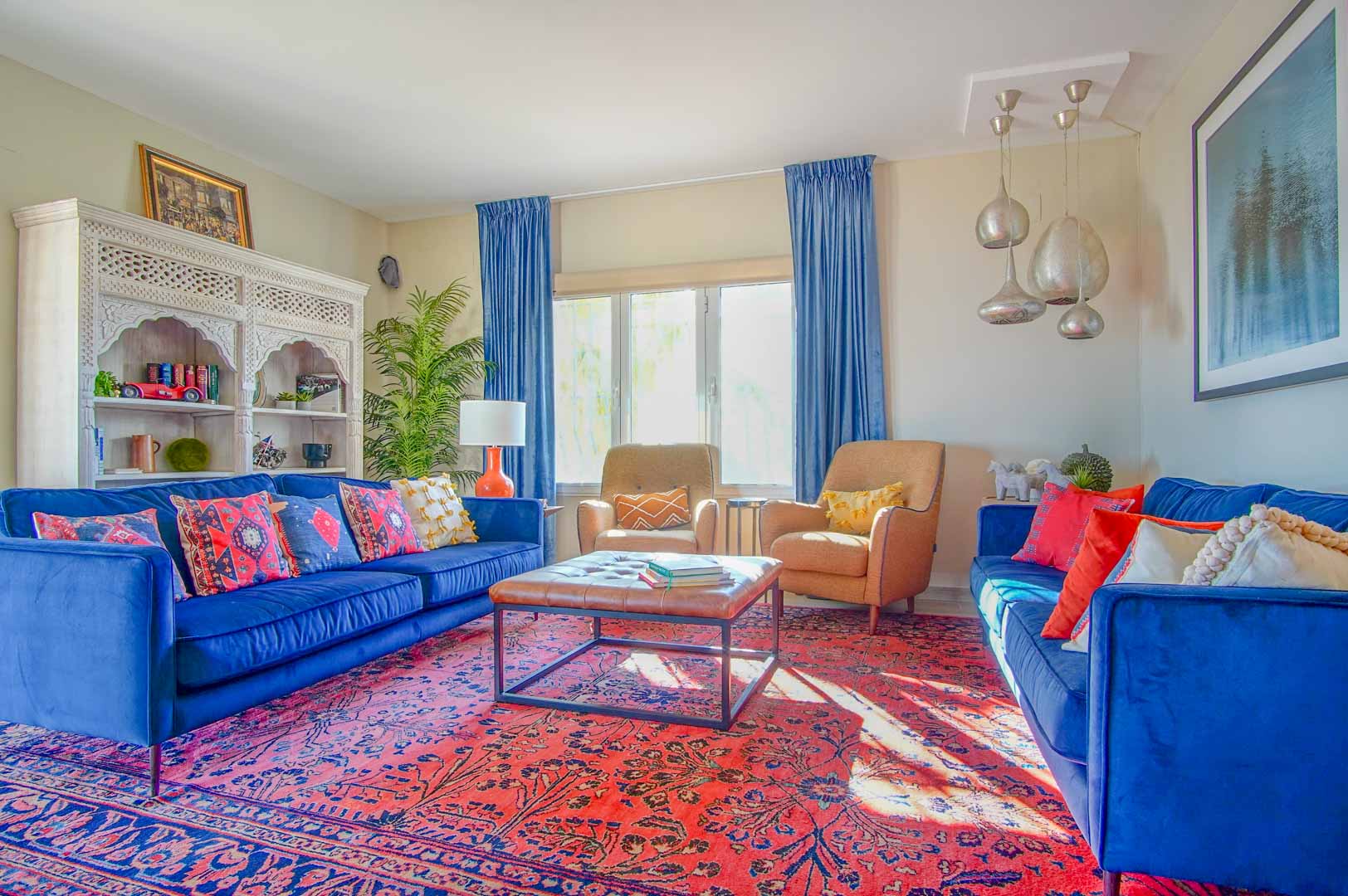Living room featuring blue velvet sofas, a red patterned rug, and a carved white bookshelf. Natural light illuminates the space, complemented by silver pendant lighting.