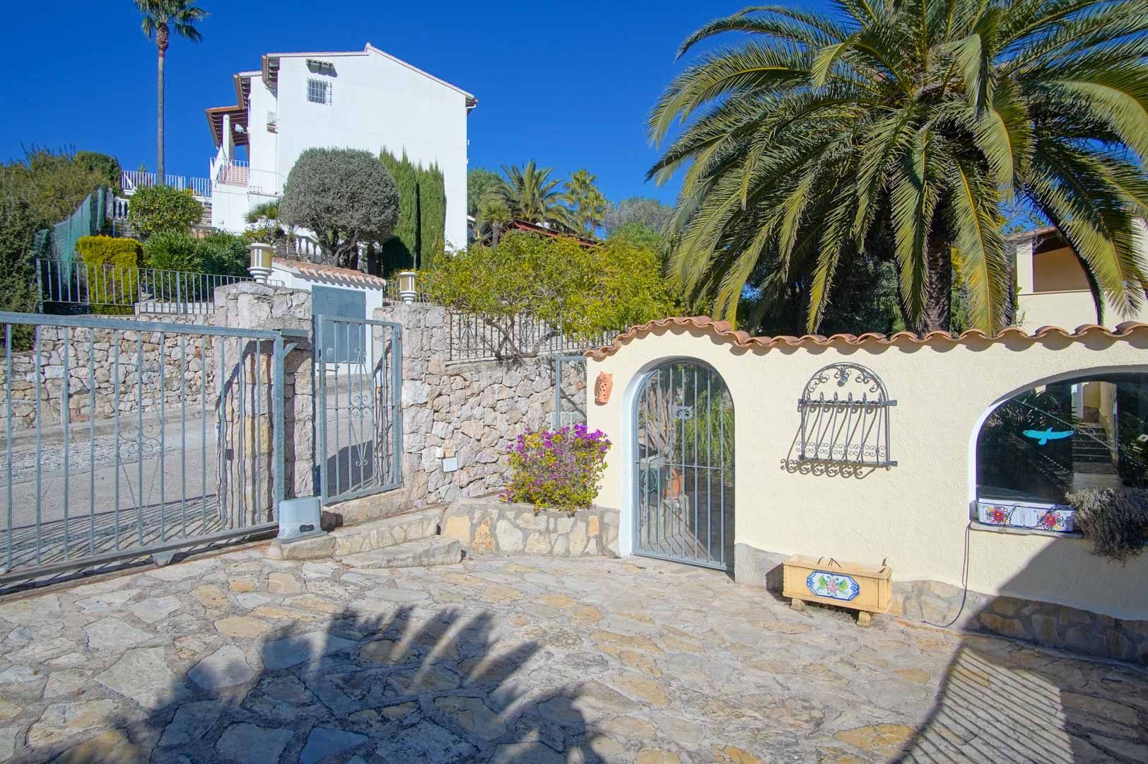Main entrance featuring a metal sliding gate, natural stone walls, and an arched pedestrian doorway leading to the villa's paved courtyard.
