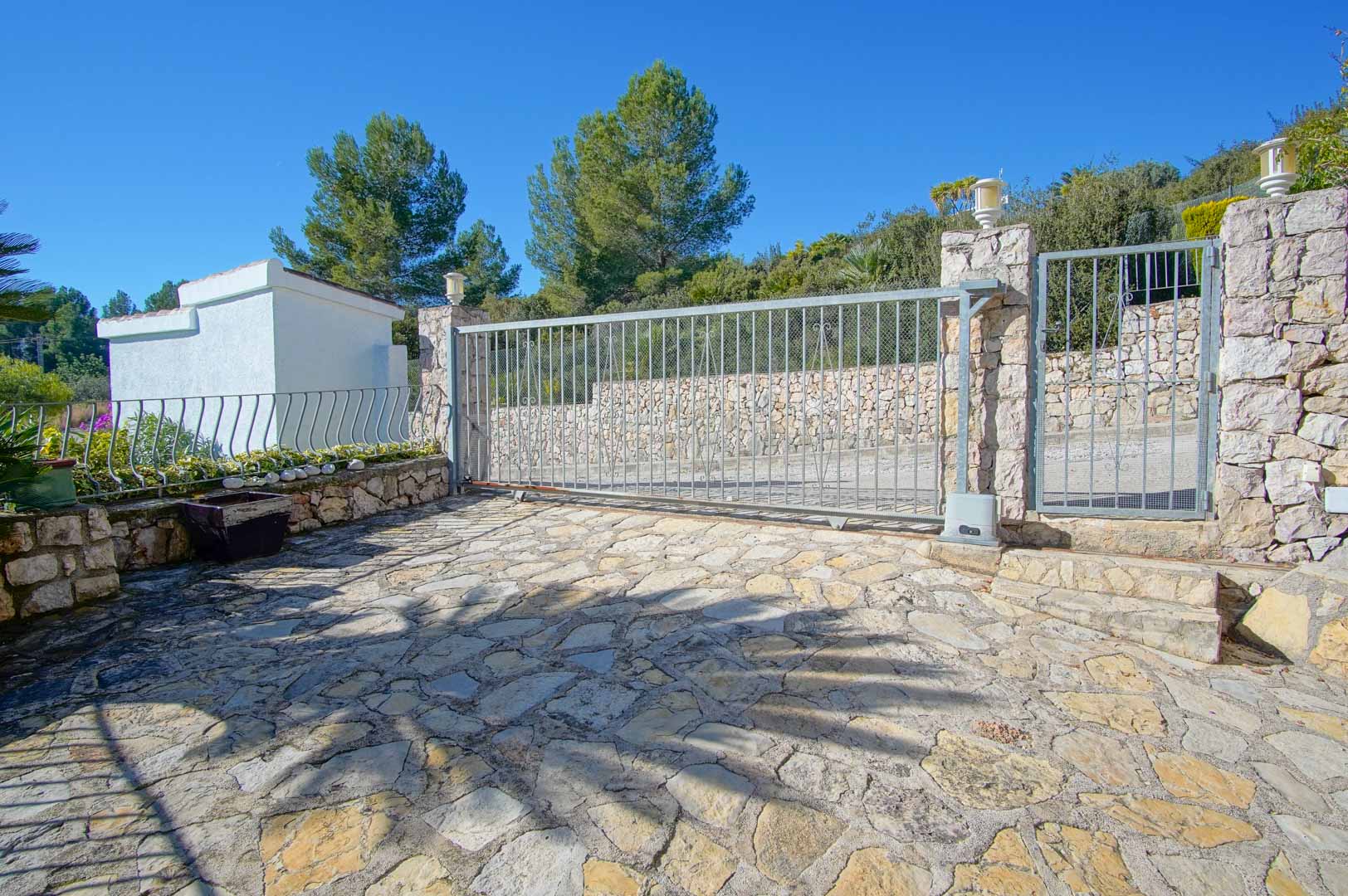 Main entrance featuring an automated sliding gate and pedestrian door, framed by natural stone walls and irregular flagstone paving under a clear sky.