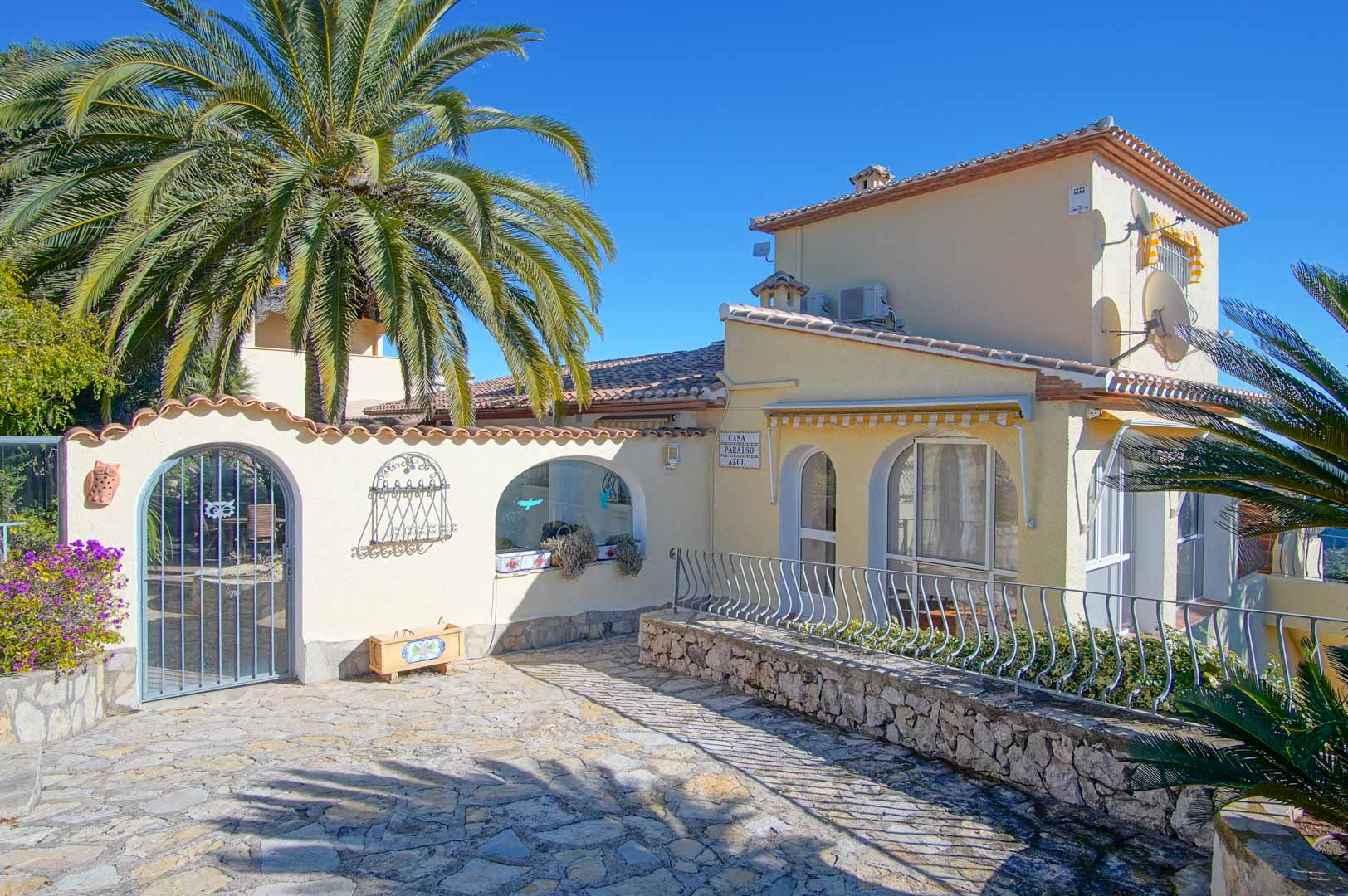 Main facade of the villa featuring a stone-paved entrance, natural stone walls, decorative arches, and Mediterranean palm trees under a clear sky.