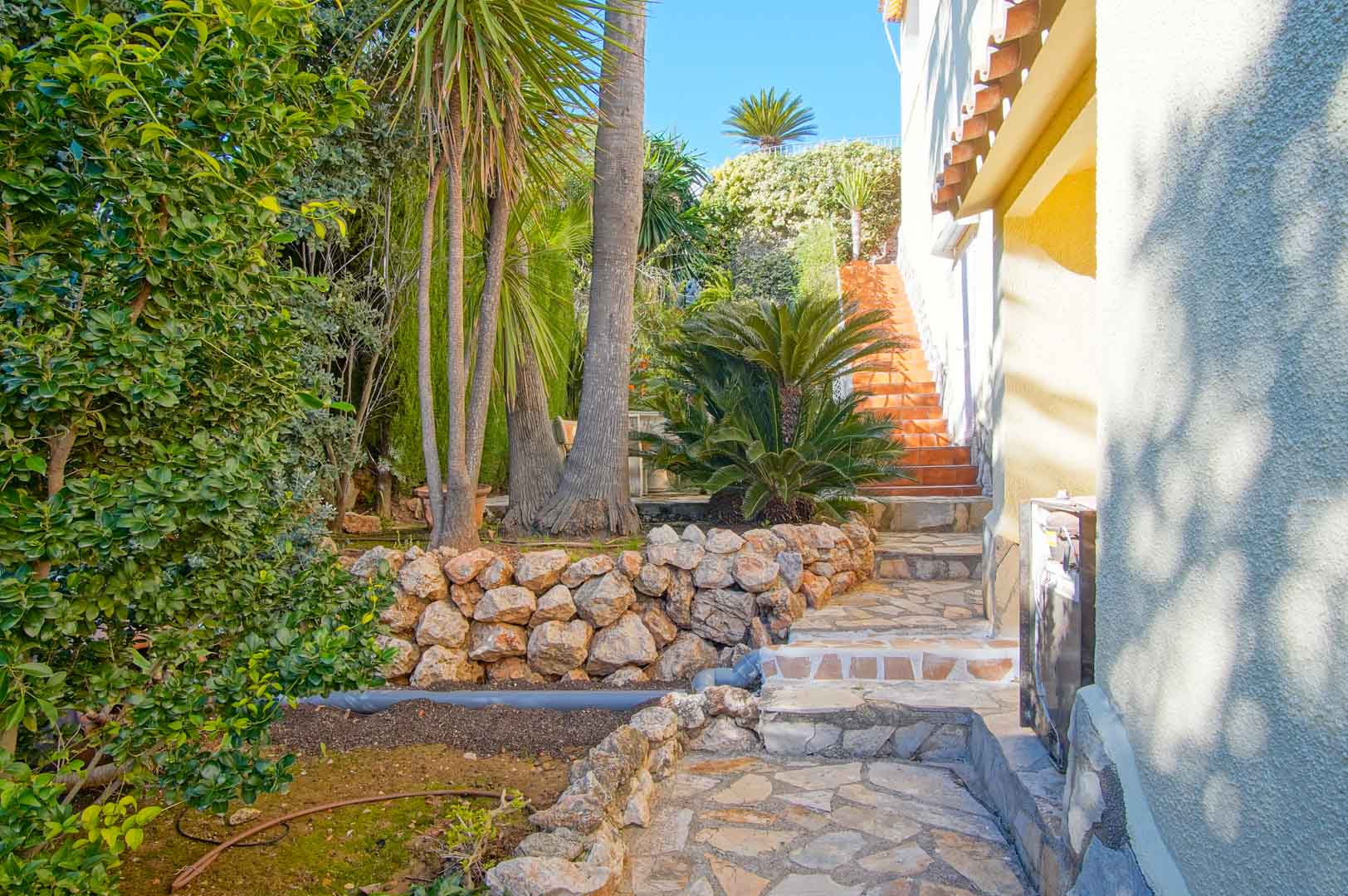 Natural stone pathway flanked by dry stone walls and lush Mediterranean flora, leading to terracotta-tiled stairs alongside the villa's white textured facade.