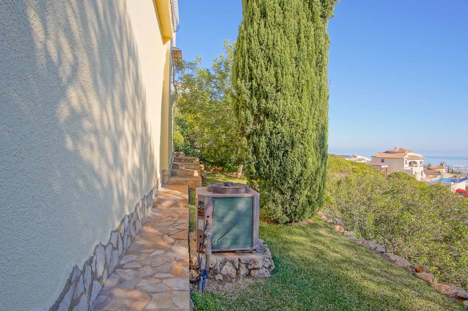 Natural stone side path along the textured facade, offering distant sea views and mature Mediterranean landscaping with a prominent cypress tree.