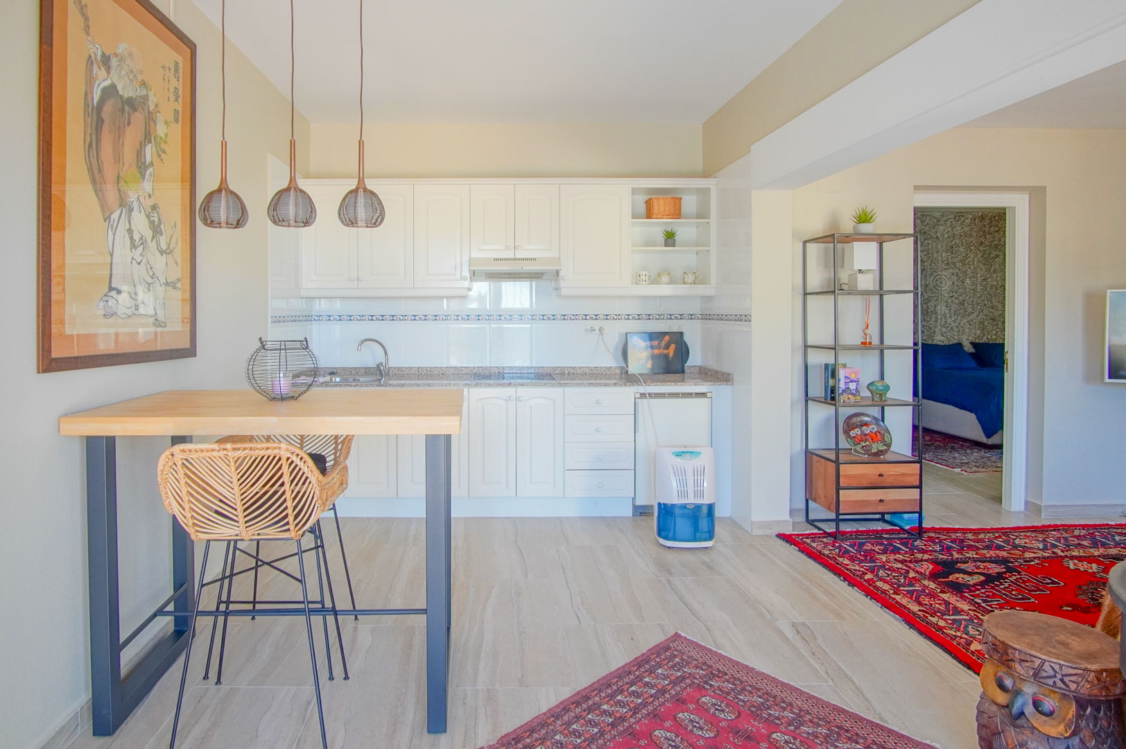 Open-plan kitchen featuring white cabinetry, granite countertops, and a wooden breakfast bar with wicker seating. The area flows into a living space with traditional rugs.