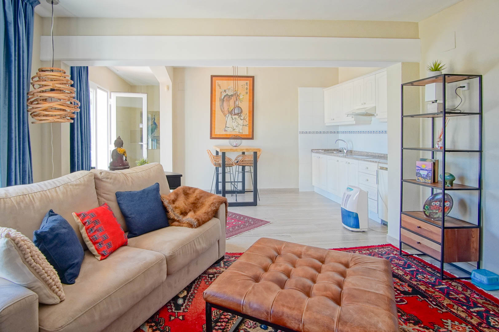 Open-plan living area featuring a compact white kitchenette, leather ottoman, and dining nook, illuminated by natural light from the terrace door.