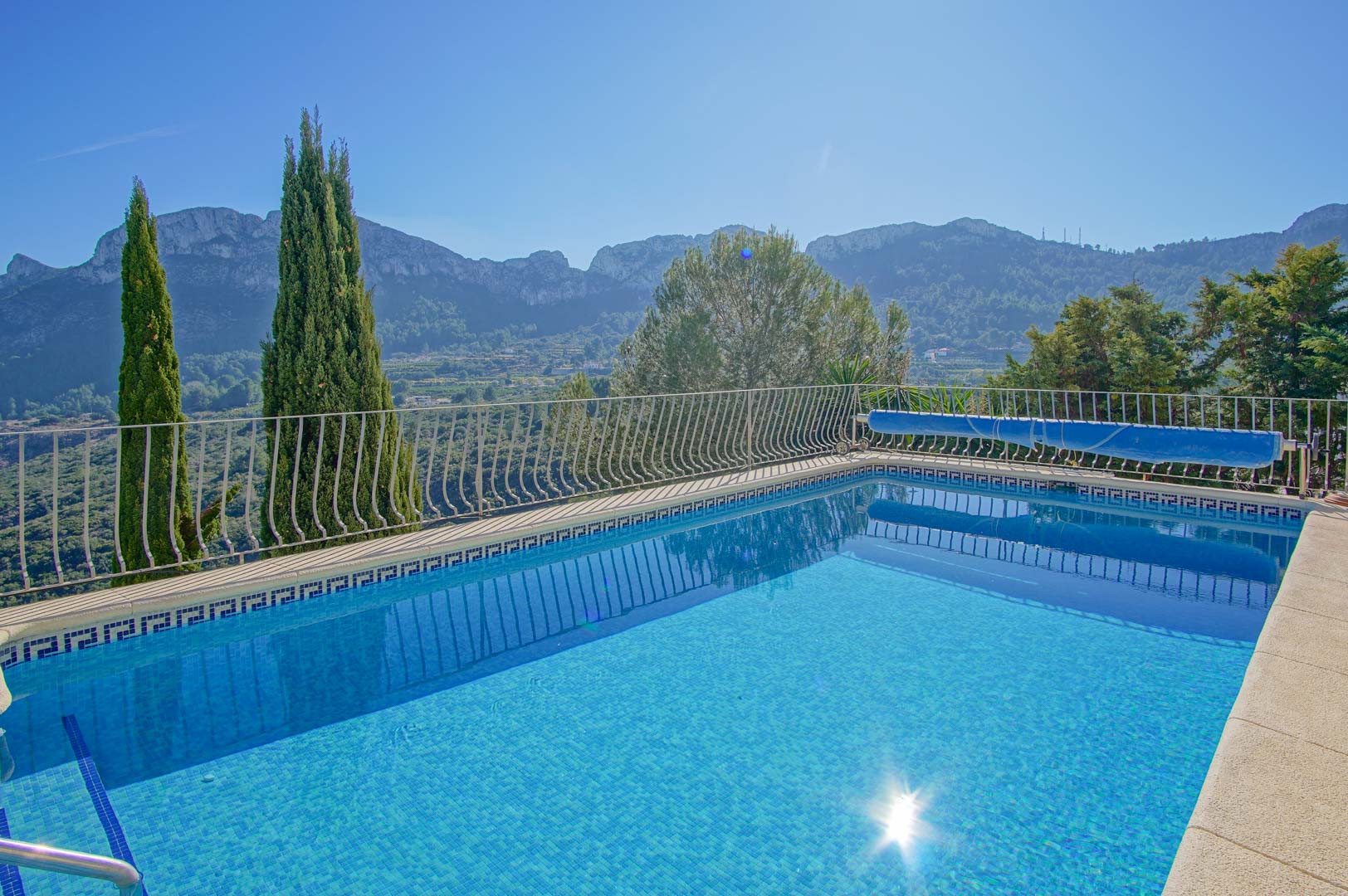 Outdoor swimming pool featuring blue mosaic tiling, a safety railing, and expansive views of the surrounding mountains and Mediterranean landscape.