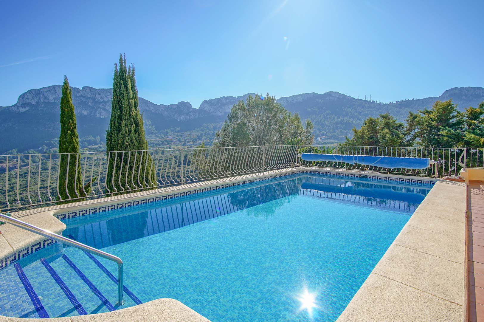 Outdoor swimming pool featuring blue mosaic tiling, integrated steps with a handrail, and a perimeter railing overlooking the mountain landscape.