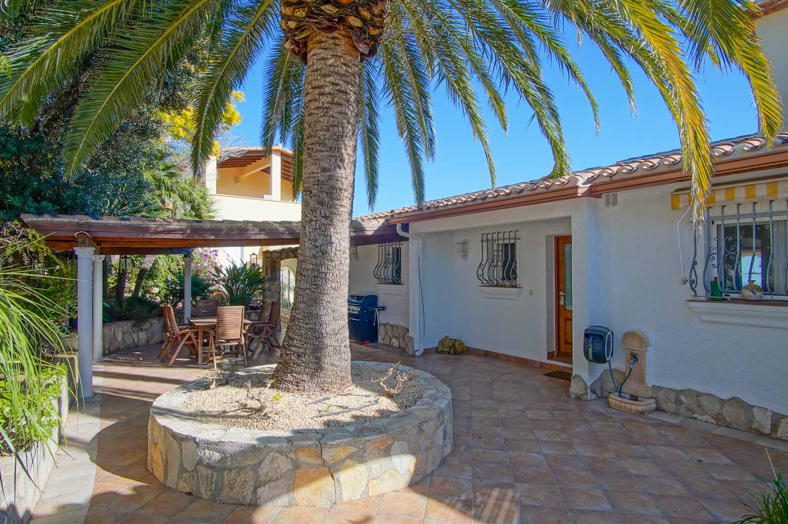 Outdoor terrace featuring a central palm tree in a stone planter, terracotta tiling, and a shaded wooden pergola with a dining set.