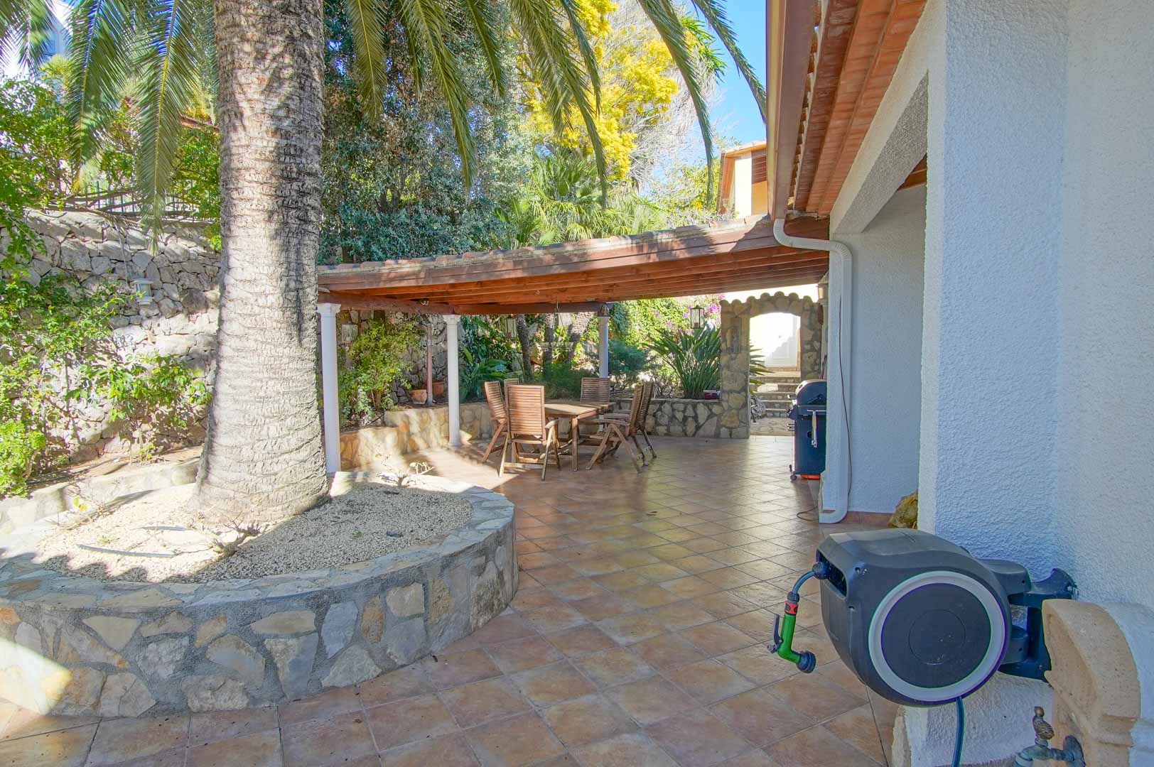 Outdoor terrace featuring a wooden pergola-covered dining area, natural stone walls, and a large palm tree set within a tiled patio.