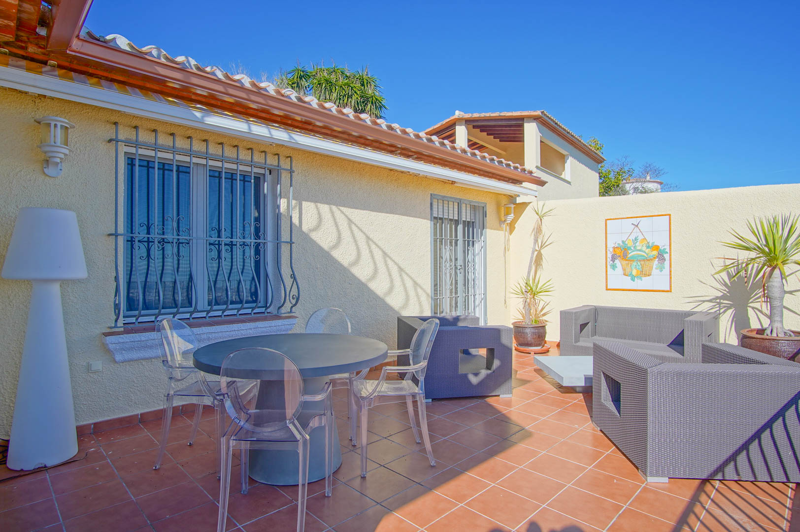 Outdoor terrace featuring terracotta tiling, a round dining table with transparent chairs, and a grey wicker lounge set against a textured facade.