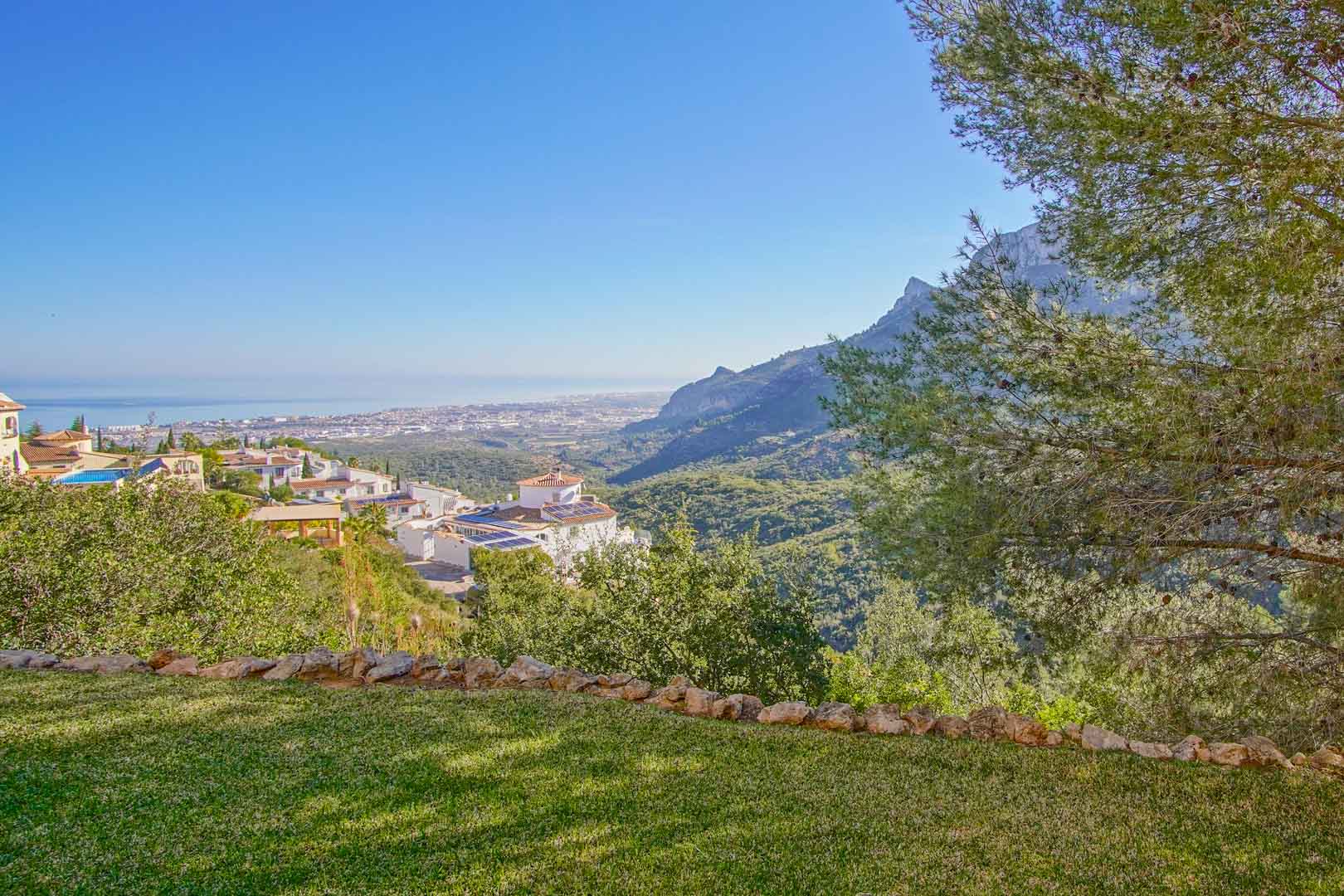Panoramic views from the manicured lawn overlooking the Mediterranean coast and rugged mountain ridges, framed by mature pine trees and greenery.