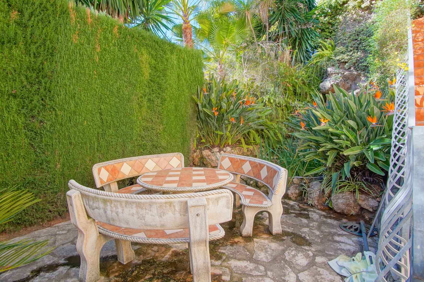 Secluded outdoor seating area featuring a stone table and curved benches with tile inlays, nestled among lush Mediterranean plants and Bird of Paradise flowers.
