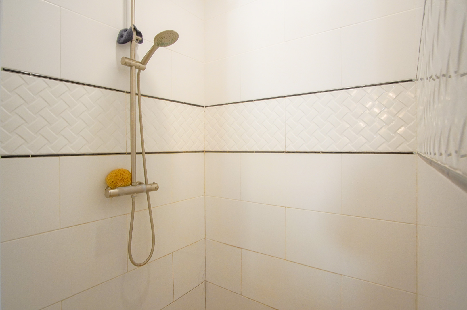 Shower area featuring white wall tiles, a textured basket-weave decorative border, and a modern stainless steel adjustable shower head.