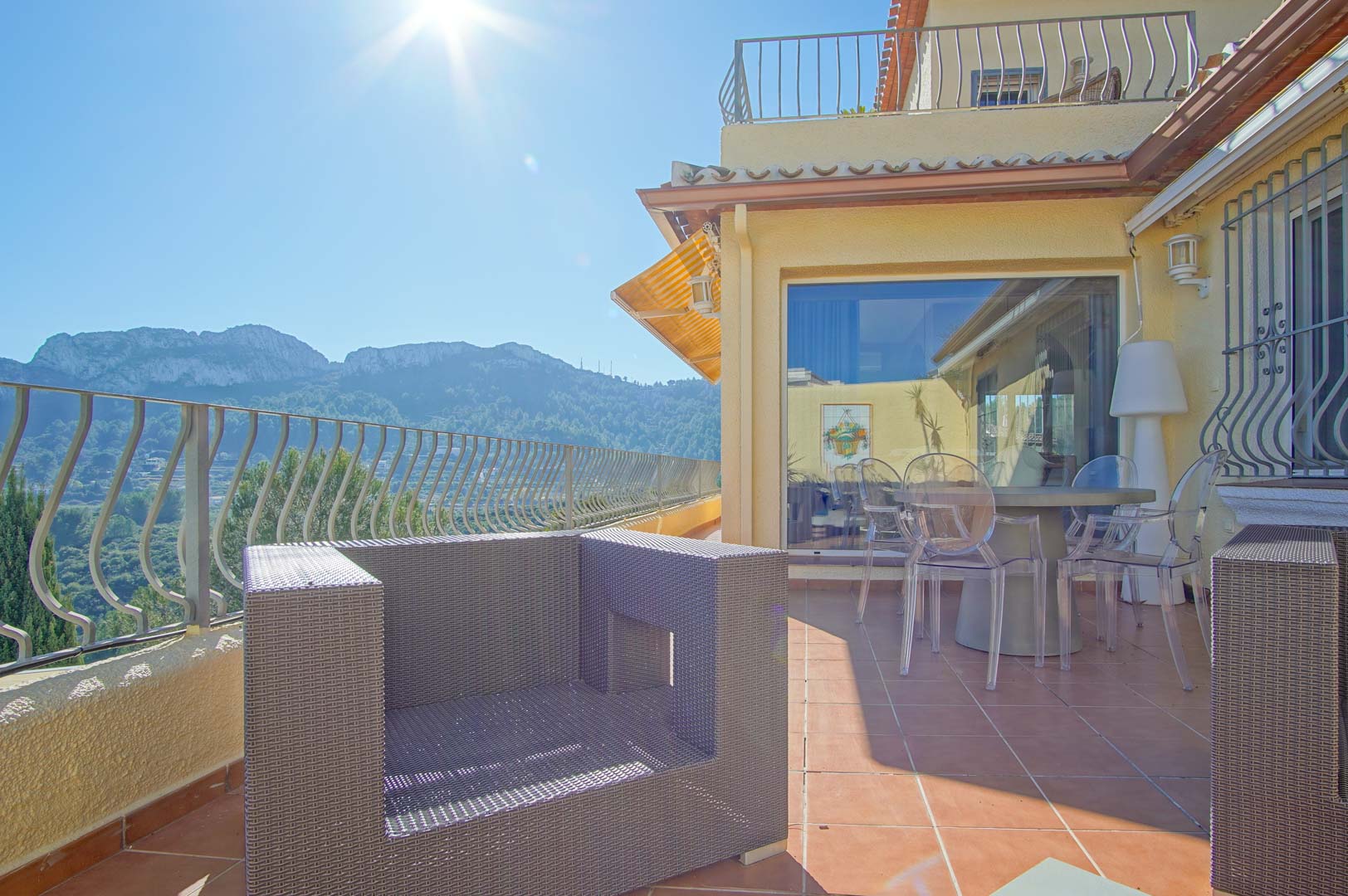 Terrace featuring terracotta tiling, synthetic rattan seating, and transparent chairs, overlooking a panoramic mountain landscape under bright sunlight.