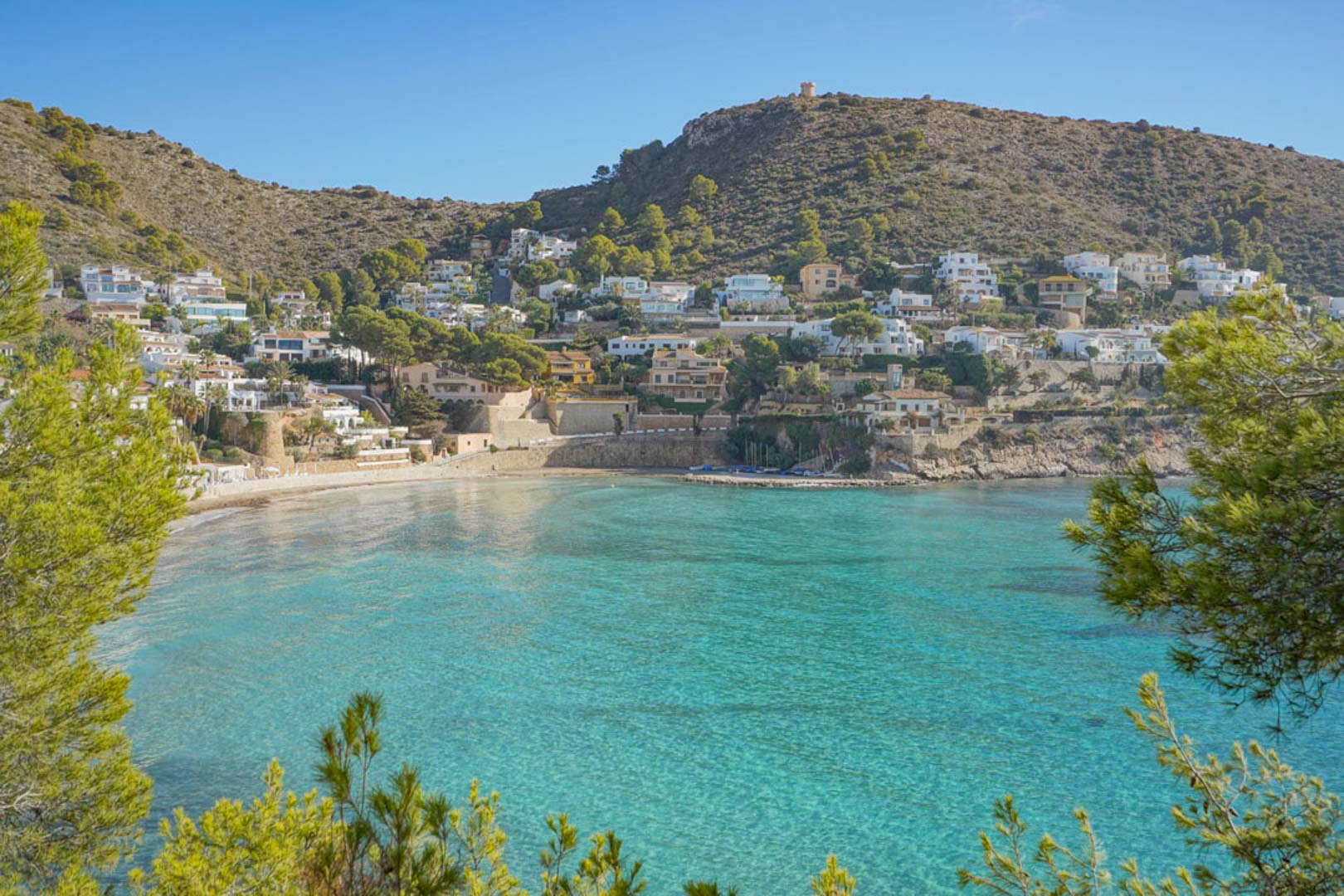 Panoramic view of El Portet bay in Moraira, featuring turquoise waters, a sandy shoreline, and Mediterranean pines overlooking the coastal residential area.