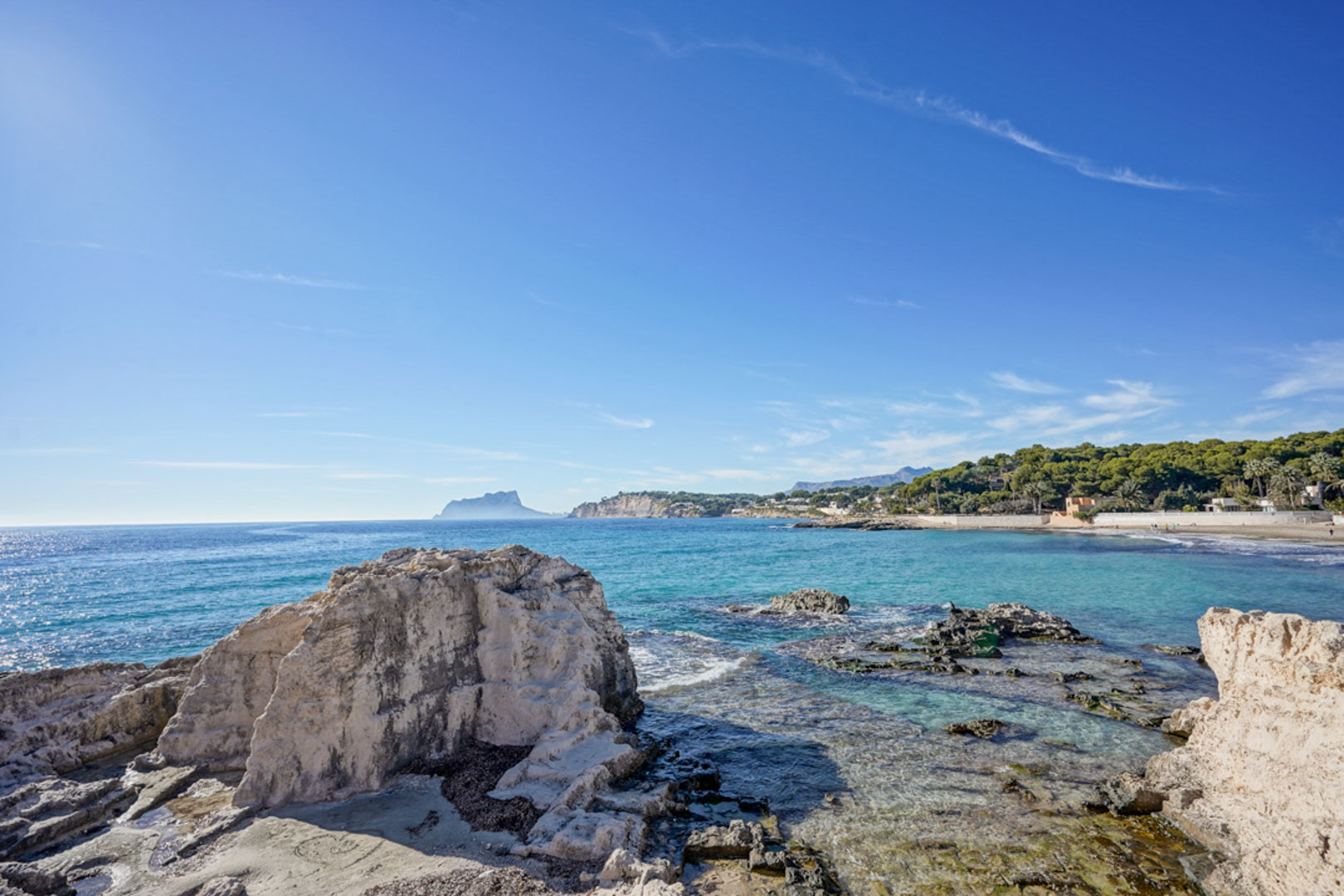 Panoramic view of the Moraira coastline featuring natural rock formations, crystal-clear turquoise waters, and the Peñón de Ifach landmark on the horizon.