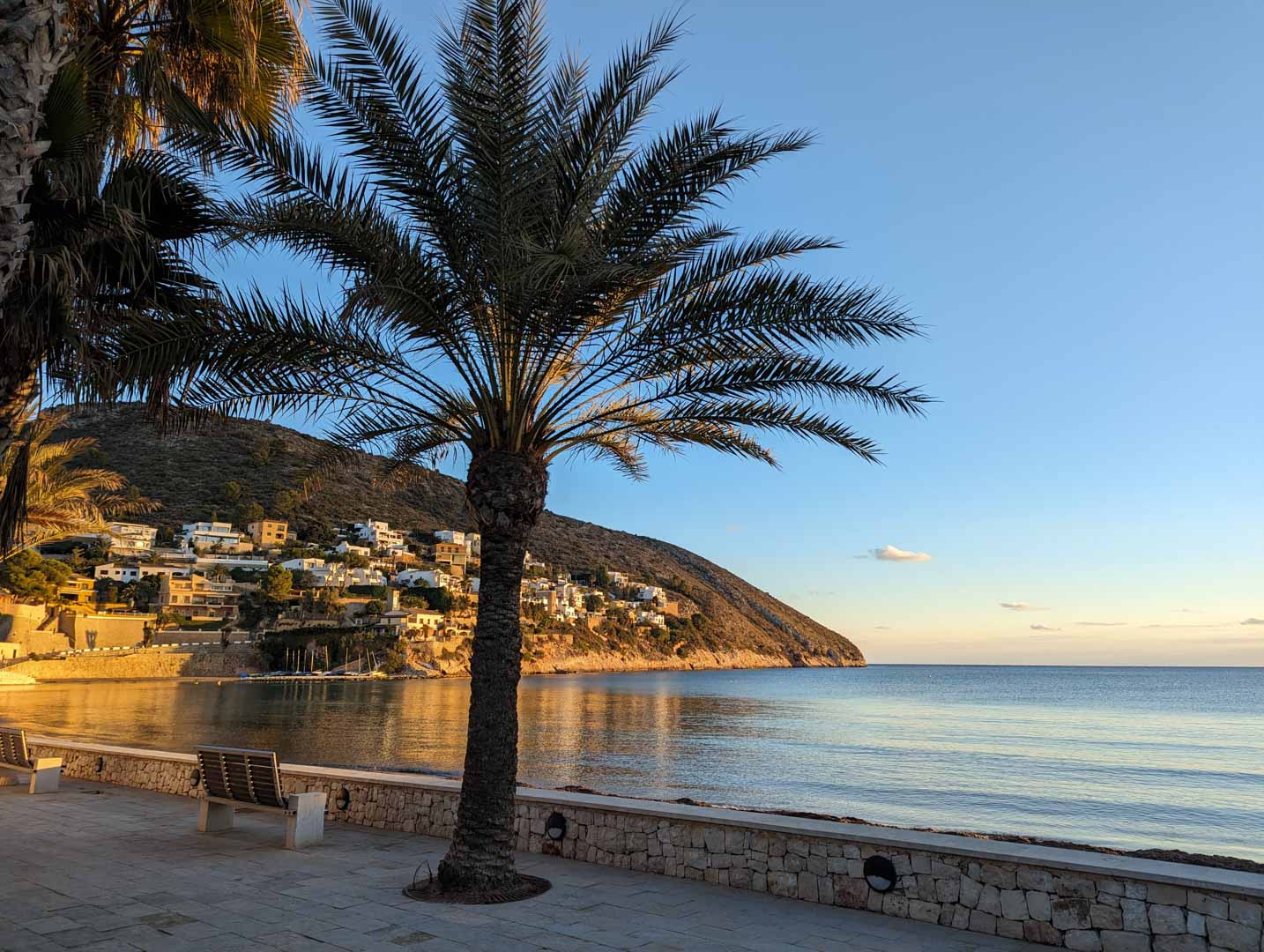 Stone-paved coastal promenade in Moraira featuring palm trees and wooden benches, with views of the calm Mediterranean Sea and hillside villas at sunset.