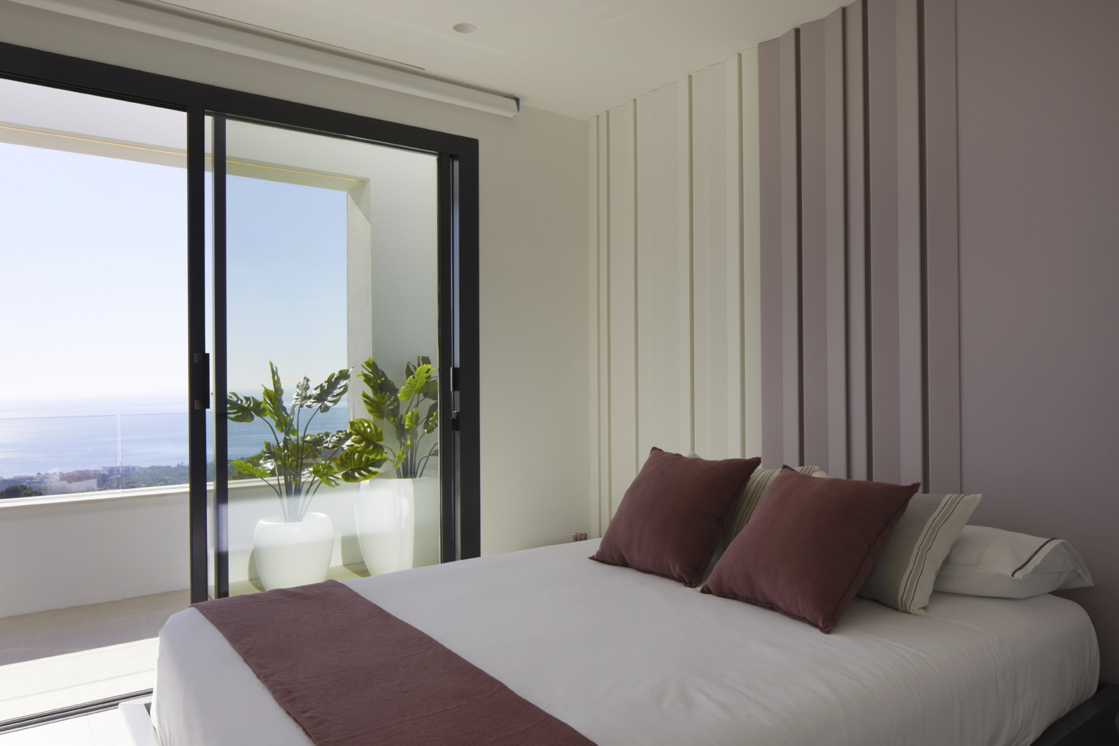 Bedroom featuring a textured vertical slat wall and sliding glass doors opening to a terrace with potted plants and expansive Mediterranean Sea views.