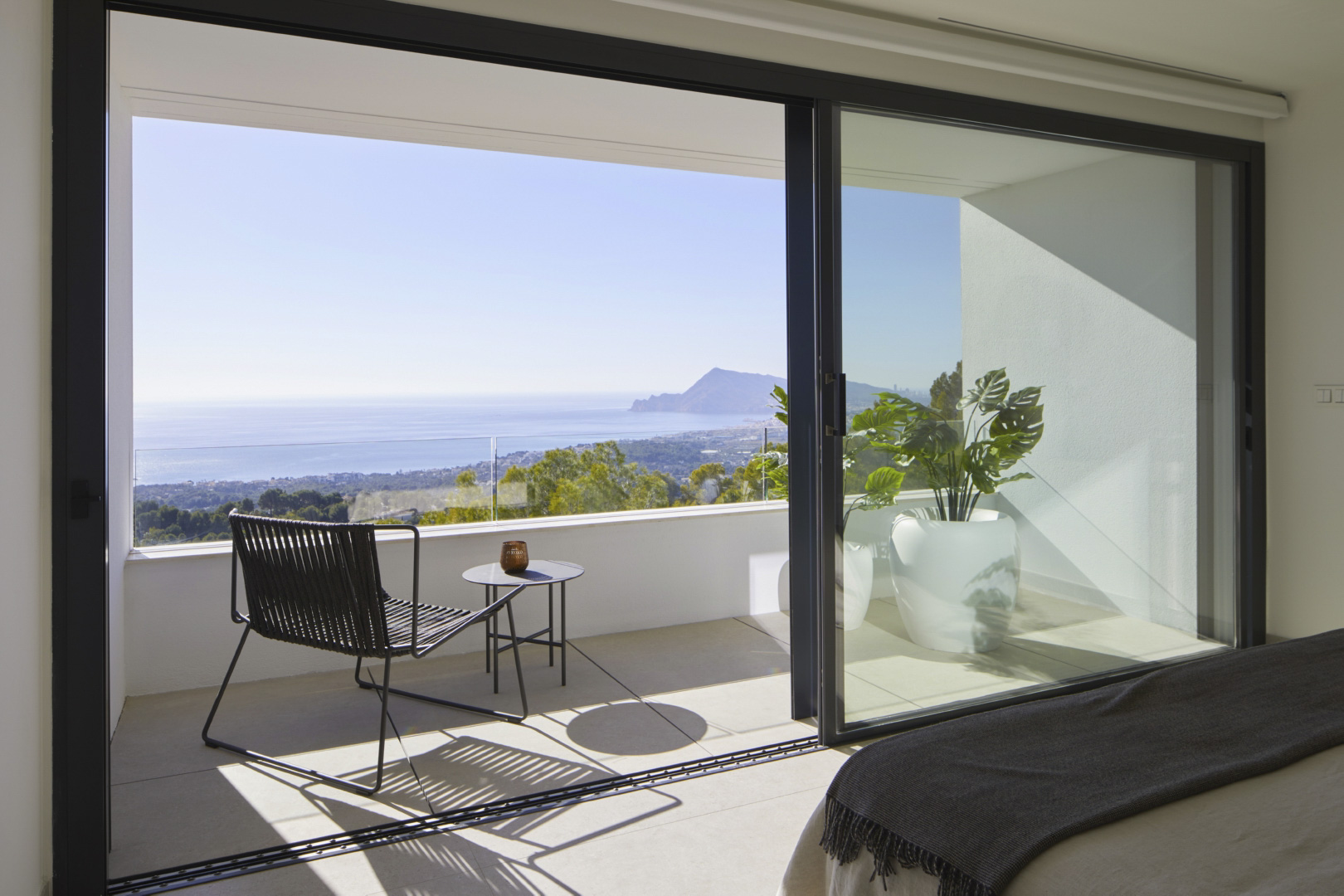 Bedroom featuring large sliding glass doors opening onto a private terrace with a glass balustrade, modern outdoor seating, and panoramic Mediterranean views.