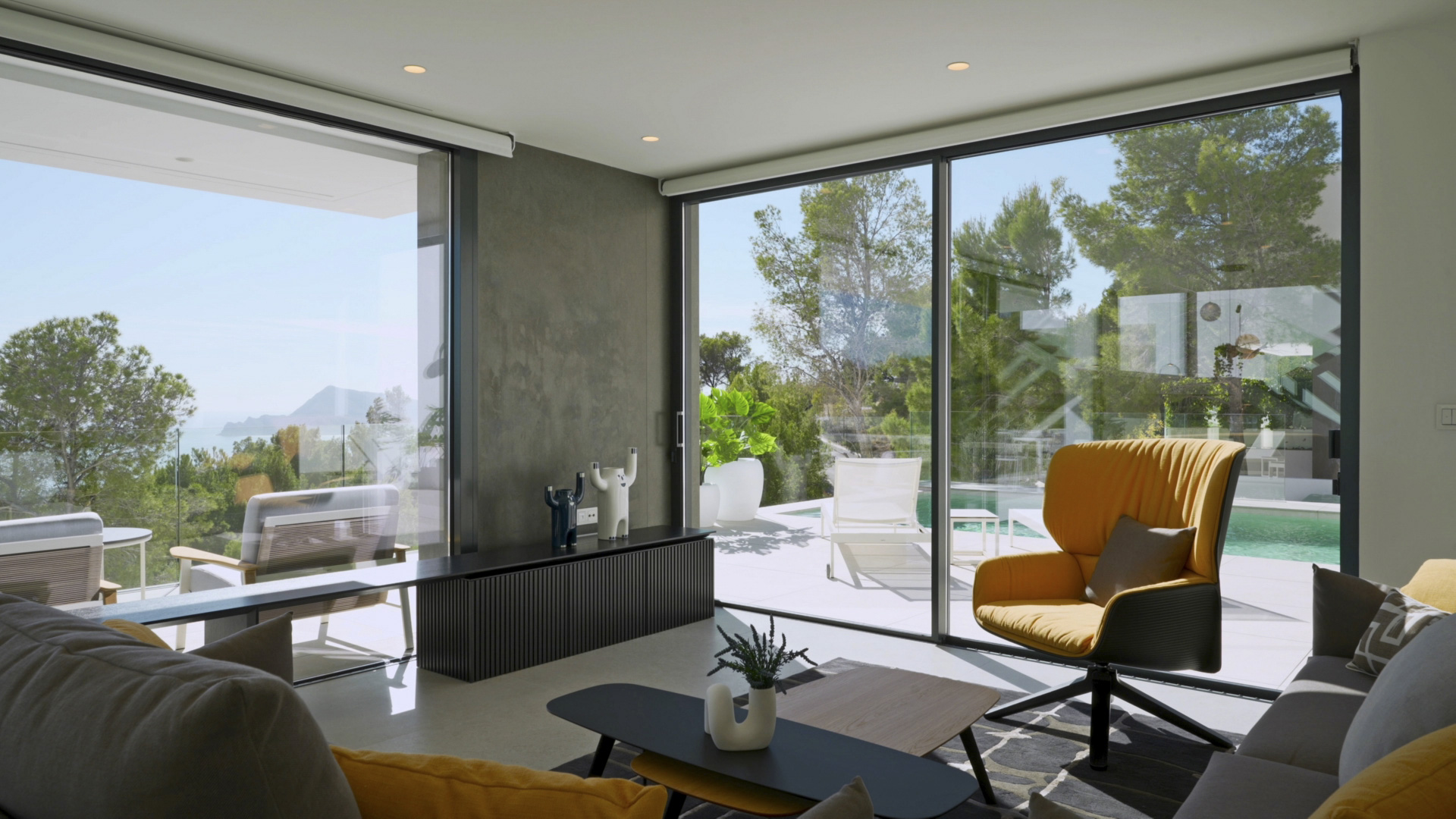 Bright living area featuring floor-to-ceiling glass doors with sea and pool views. It includes designer furniture, a mustard armchair, and direct terrace access.