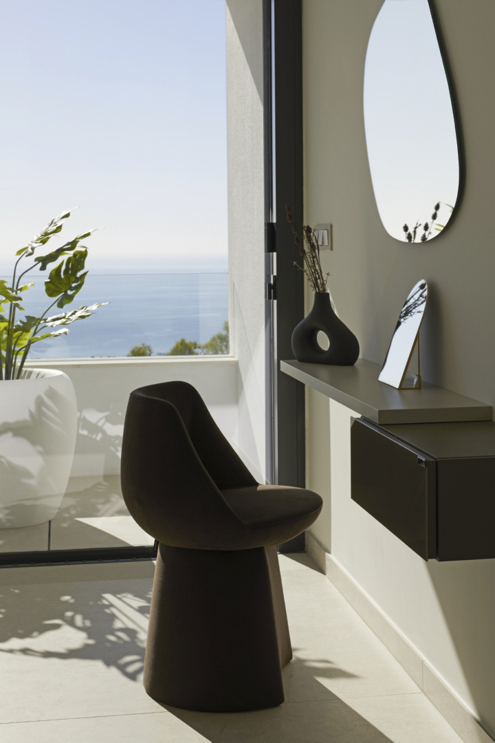 Designer dressing area featuring an organic-shaped mirror and sculptural chair, positioned by floor-to-ceiling windows with Mediterranean views.